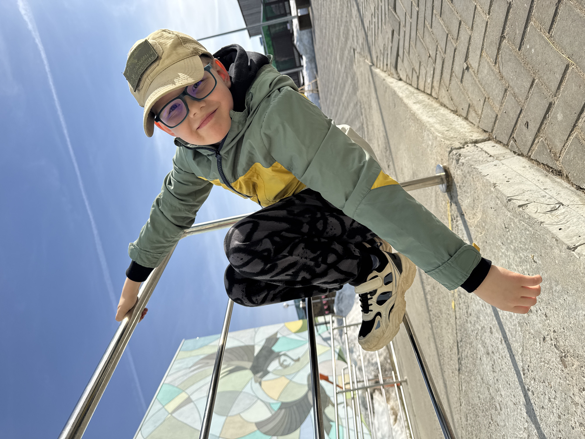 A young boy in a green and yellow jacket, black and white pants, and a tan cap with a patch and glasses is perched on a silver metal railing, leaning towards the camera. He's holding the railing with one hand, his other hand extending toward the right. Behind him is a clear blue sky. A brick pathway, a mural, and parts of a building can also be seen in the background. This image was taken in Perm, Russia.