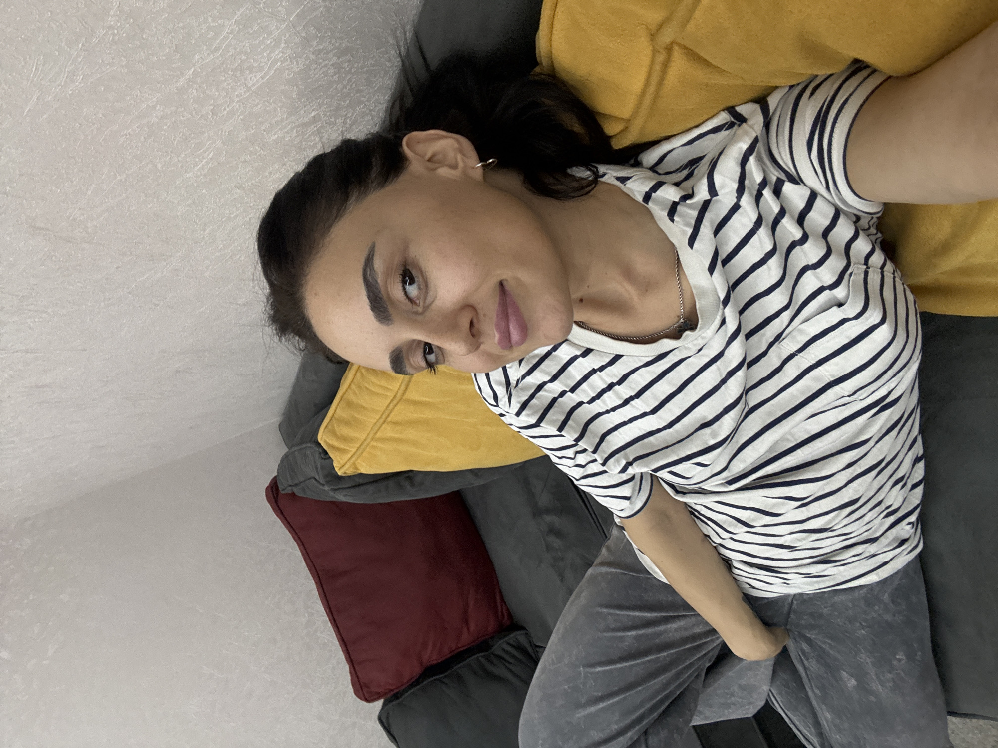 This media file shows a medium shot of a young woman lying on a sofa with her head propped up on a yellow pillow. She is wearing a black and white striped t-shirt and grey sweatpants. Her hair is dark and pulled back. She has subtle makeup on and is looking towards the camera with a relaxed expression. The sofa is dark grey, and there is a burgundy pillow visible beneath the yellow one. The background is a light-colored textured wall. The scene appears to be indoors, likely in a living room. There is no visible text in the image. The lighting suggests it could be daytime, possibly in the afternoon. The overall impression is casual and comfortable, suggesting the woman is relaxing at home in Tbilisi, Georgia.