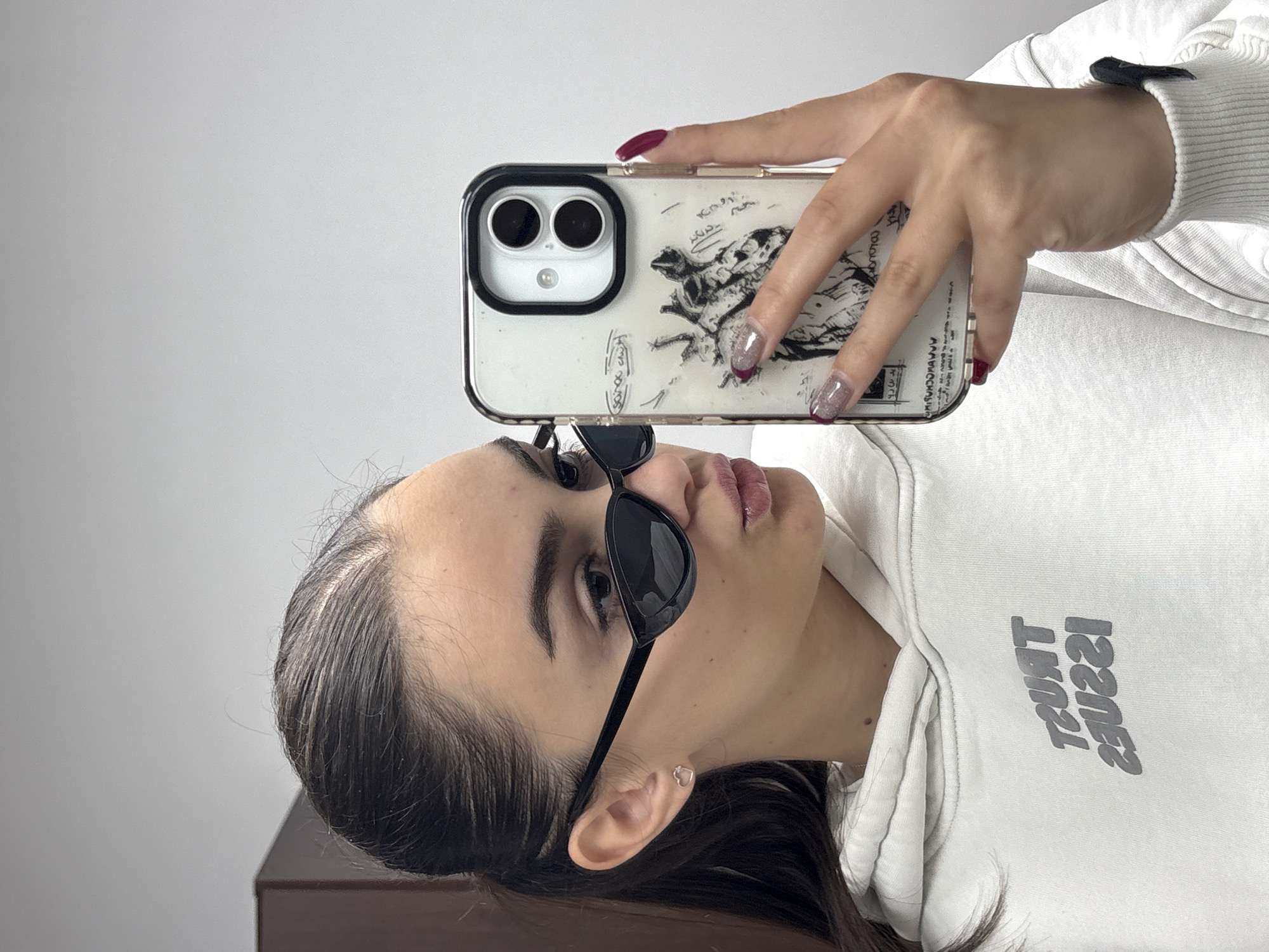 A young woman is taking a selfie with her phone. She is wearing sunglasses, a white hoodie, and earrings. Her nails are painted. The phone has a clear case with a drawing on it. The background is a plain white wall. The scene is indoors.