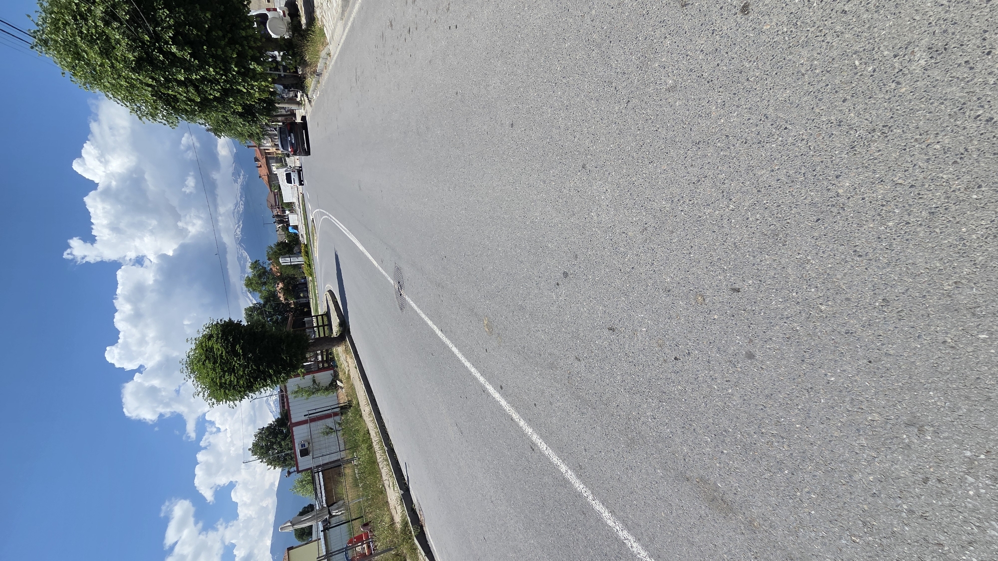 The image depicts a paved asphalt road within an unknown town or village, viewed on a clear, sunny day with a bright blue sky and scattered large, white cumulus clouds. The road extends from the foreground, gently curving towards the right in the mid-distance. A solid white line delineates the left edge of the visible lane, and a circular manhole cover is present in the asphalt near the center of this lane.
On the left side of the road, a low metal fence borders a grassy area with weeds. Behind the fence are several low-rise structures, possibly sheds or storage units, featuring corrugated metal roofs and walls of white and red siding. A single, well-maintained deciduous tree with a dense, rounded green canopy stands by the road on this side. A red object, resembling a child's toy, is visible on the ground near the fence in the lower left quadrant.
On the right side of the road, a line of mature deciduous trees with abundant green foliage runs parallel to the street. Further along, residential buildings with reddish-brown tiled roofs are visible. In the distance, a white van and a dark-colored sedan are either parked or driving on the road. A white monument or statue is partially visible beyond these vehicles on the right.
The background is dominated by a range of majestic snow-capped mountains, suggesting a mountainous geographical location for the depicted settlement. No individuals are visible in the immediate scene.