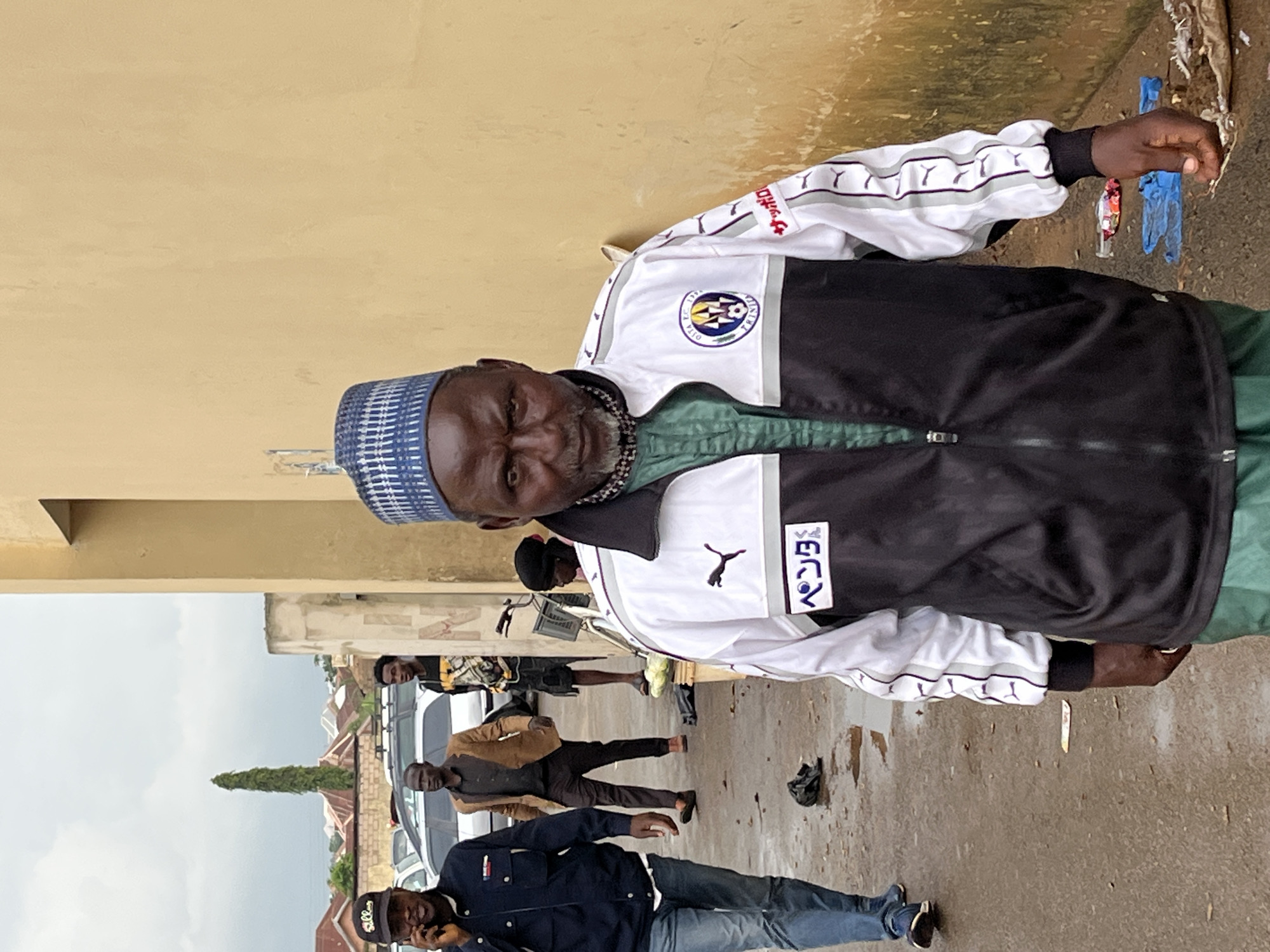 In Bukuru, Nigeria, an elderly man wearing a blue and white patterned cap and a Puma tracksuit is the focal point of the image. The man has dark skin, a full beard, and is looking directly at the camera with a serious expression. He is wearing a green shirt beneath his tracksuit jacket, which features a Puma logo on the left chest and Japanese text on the right side of the chest. The tracksuit also has white stripes with the Puma logo repeated along the sleeves. He is standing on a wet, paved surface, possibly a street or walkway, with scattered debris including plastic bags and what appears to be dirt.
In the background, several other individuals are visible. To the left, a man wearing a dark blue jacket and jeans is walking away from the camera, holding a phone to his ear. Behind him, two other men are walking. One is wearing a brown jacket over a dark shirt and trousers, while the other is wearing a black and yellow patterned t-shirt and shorts. Several parked cars are also visible behind these individuals. A tall, slender tree stands in the distance. A woman wearing a headscarf is seated next to a bicycle in the middle ground, to the right of the parked cars. The scene appears to be in an urban or semi-urban environment with buildings visible in the distance. The overall lighting suggests an overcast day.