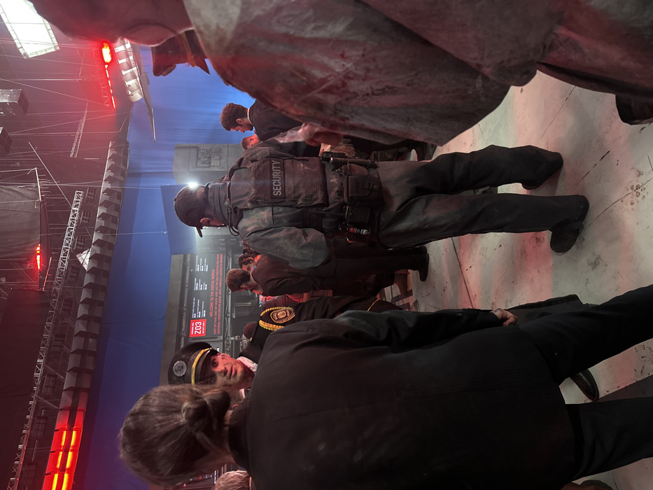 The scene depicts an interior film set within a soundstage, likely at the Qingdao Oriental Movie Capital in Huangdao District, China. The environment features a large blue screen serving as a backdrop, indicating chroma key usage. Overhead, exposed metal truss structures support numerous red stage lights and diffused white panel lights, creating a low-key, dramatic illumination. The floor is a light-colored concrete or industrial surface with visible scuff marks and debris.
In the foreground, a person with dark hair secured in a bun, wearing a dark suit jacket, stands with their back mostly to the camera. To their immediate right, partially visible, is another individual in a light-colored, distressed garment exhibiting simulated bloodstains on the right shoulder and arm.
In the midground, a male individual faces partially towards the camera, wearing a dark uniform cap with gold trim and insignia, and a dark uniform jacket with an emblem on the left sleeve. His face displays simulated grime or bloodstains, and he holds a subtle expression. To his right, another male stands with his back to the camera, wearing a dark cap, a heavy dark-grey tactical vest prominently labeled "SECURITY" in white lettering on the back, and matching distressed trousers. The vest and belt are equipped with multiple pouches and gear.
In the background, a large dark structure features illuminated panels displaying text, including "Z03" and "WELCOME TO THE DIGITAL WORLD." Several other individuals, generally dressed in dark suits or attire, are visible throughout the midground and background, standing in various positions. The collective posture suggests an assembly or waiting period within a staged environment.