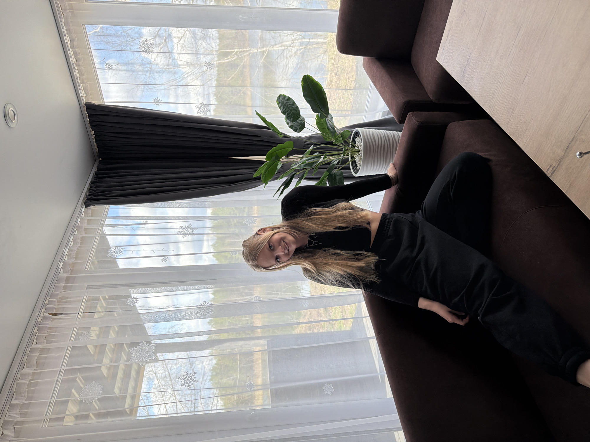 A blonde-haired, fair-skinned woman wearing a black long-sleeved shirt and black sweatpants reclines on a dark brown couch. A houseplant in a white pot sits on the couch near her head. The couch appears to be part of a sectional and has a light brown side table attached. The room has large windows covered by two layers of curtains: dark gray curtains that are mostly drawn and white, sheer curtains with snowflake designs. Outside the window, trees and a blue sky are visible. The location is likely Novyi, Russia based on image context clues.