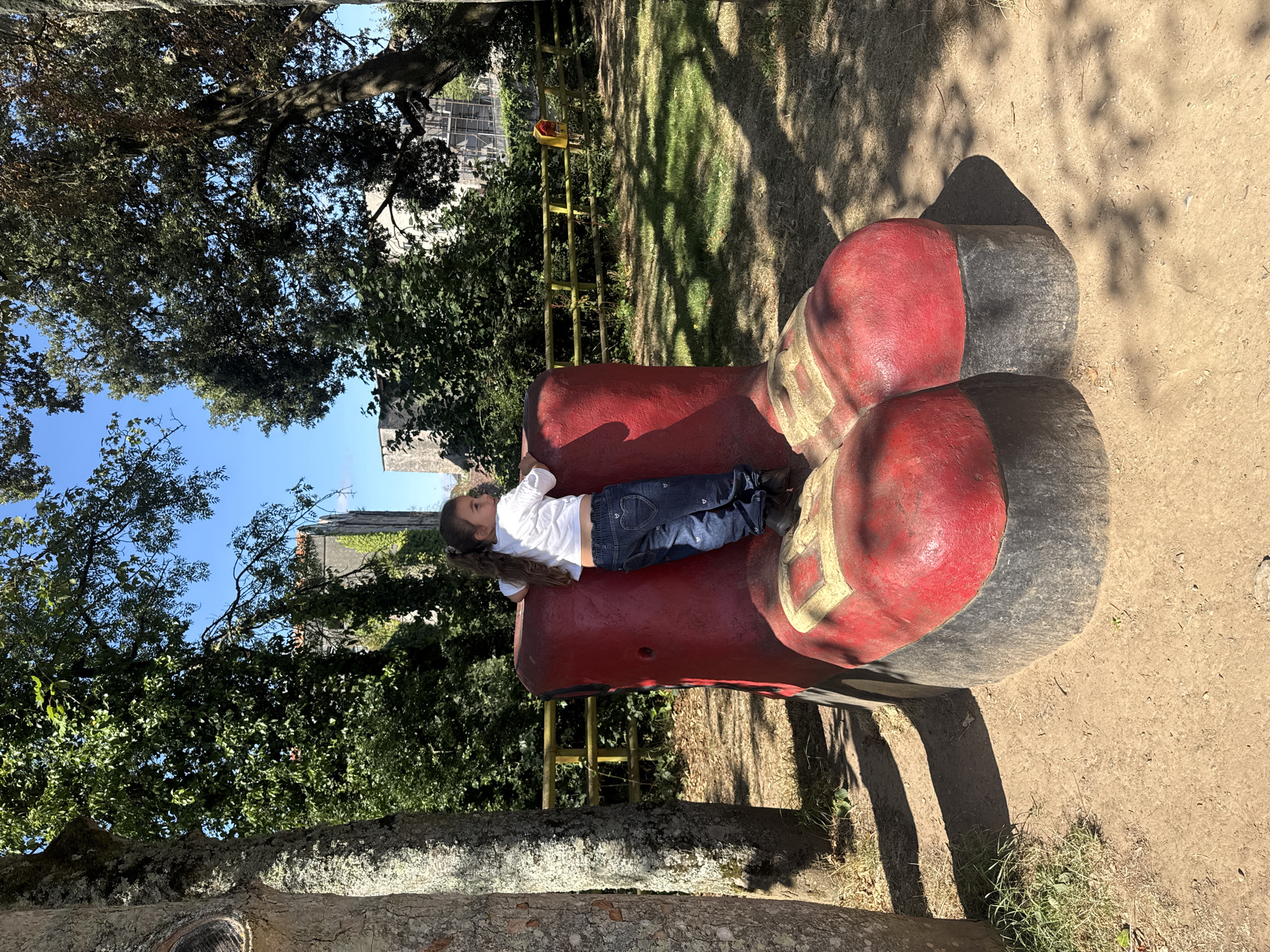 A young girl with brown hair tied in a ponytail, wearing a white t-shirt and blue jeans with heart accents, is climbing on a large, red, novelty boot sculpture. The boot has black soles and gold detailing on the toes. She is positioned on the side of the boot, looking away from the camera. The sculpture is situated outdoors on a dirt path with grass and trees in the background. Behind the trees, a stone building with ivy climbing its walls and a yellow fence are visible. The scene suggests an outdoor recreational area, potentially a park or playground, within the grounds of Cahir Castle in Cahir, Ireland, given the architectural style and surrounding landscape. The lighting indicates it is daytime, likely sunny, with shadows cast by trees.
