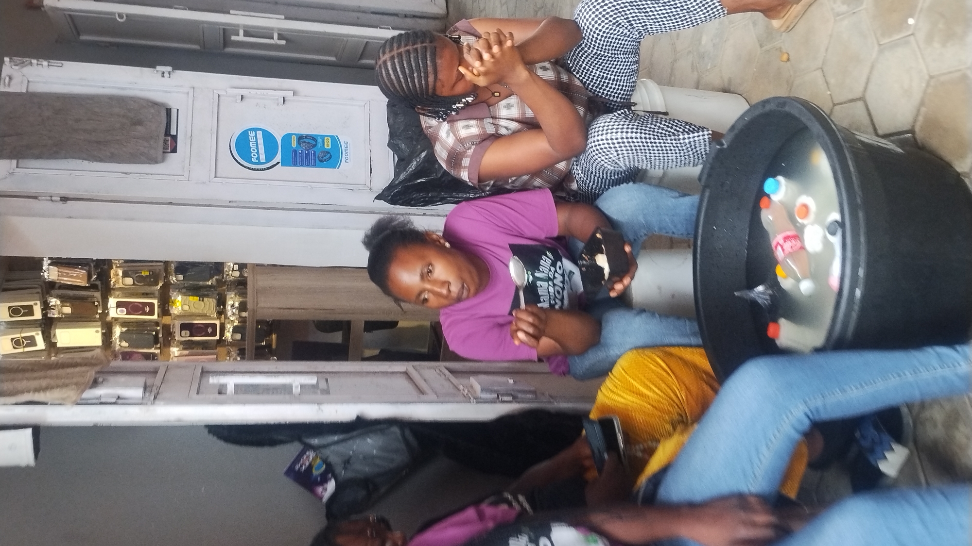 Two women are sitting outdoors, one with hands clasped in front, wearing a plaid shirt and checkered pants; the other woman is wearing a pink t-shirt and blue jeans, holding a spoon and a dark bowl. Between them is a black plastic tub filled with water and several bottles. A third person's legs and yellow fabric are partially visible near the tub. In the background is a display case with cell phone cases. The scene is located in Jos, Nigeria.