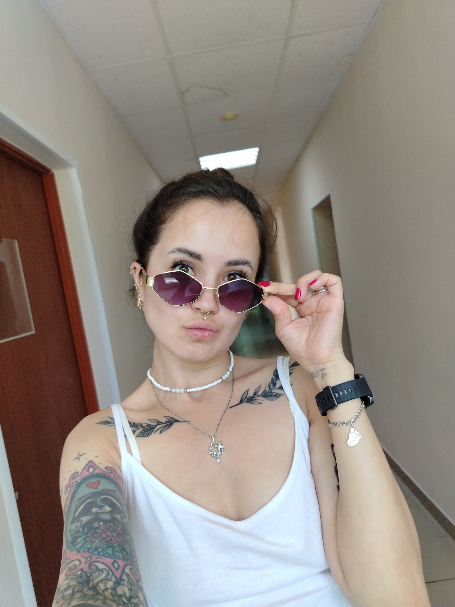 A young woman is taking a selfie in a hallway. She has dark hair pulled back in a bun and is wearing a white tank top, sunglasses, and several necklaces. She is also wearing a watch and a bracelet. The hallway appears to be in a residential building, with a door visible on the left side of the frame. The woman is looking at the camera with a playful expression.