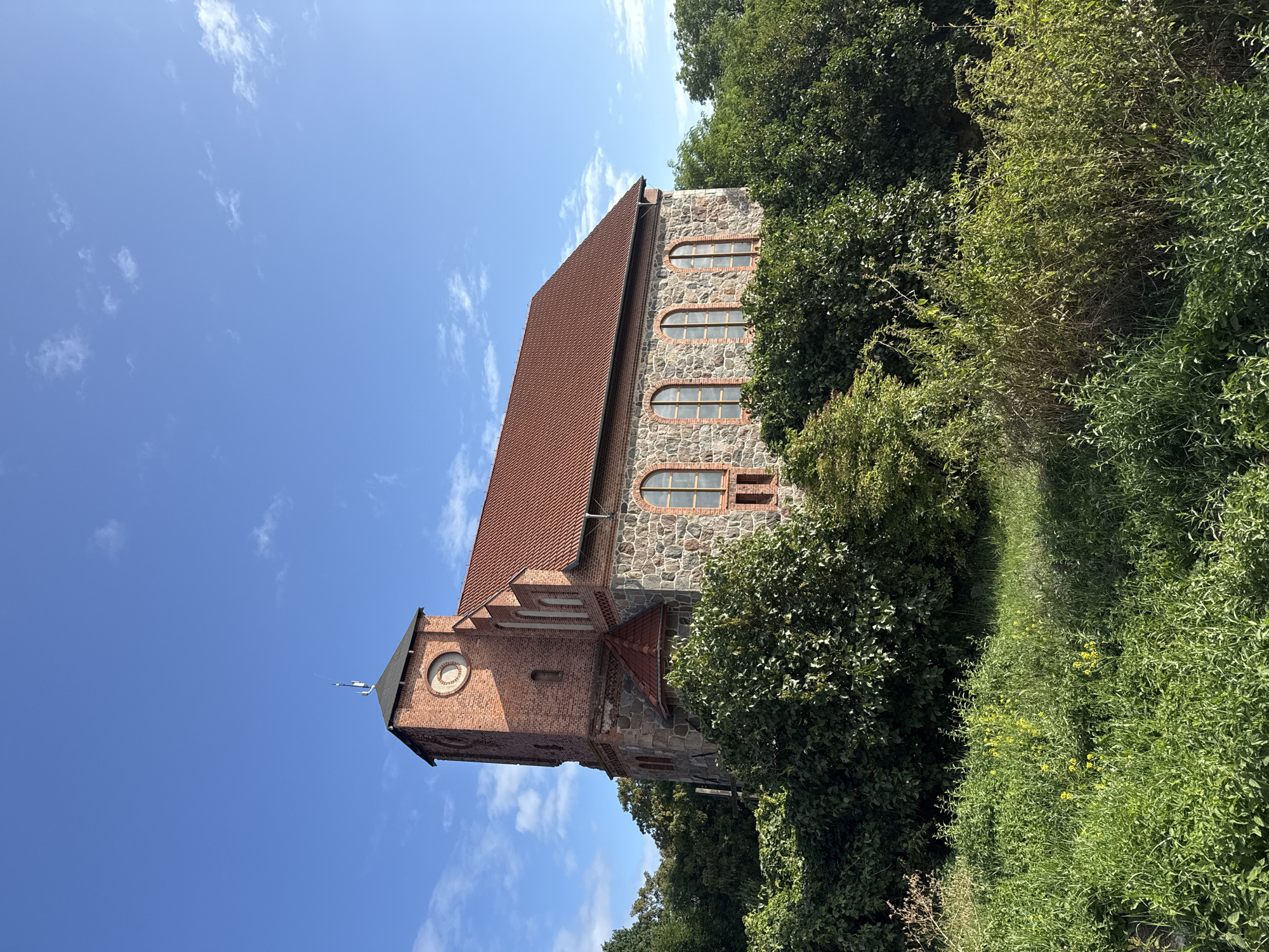 A stone and brick church with a red tile roof stands on a hill, surrounded by lush green trees and grass. The church has a prominent brick tower with a clock face. The windows are arched, and the building is constructed with a mix of stone and brick. The sky is blue with a few scattered white clouds. In the foreground, there is dense vegetation and some yellow wildflowers. The scene appears to be a sunny day in Stare Brynki, Poland.
