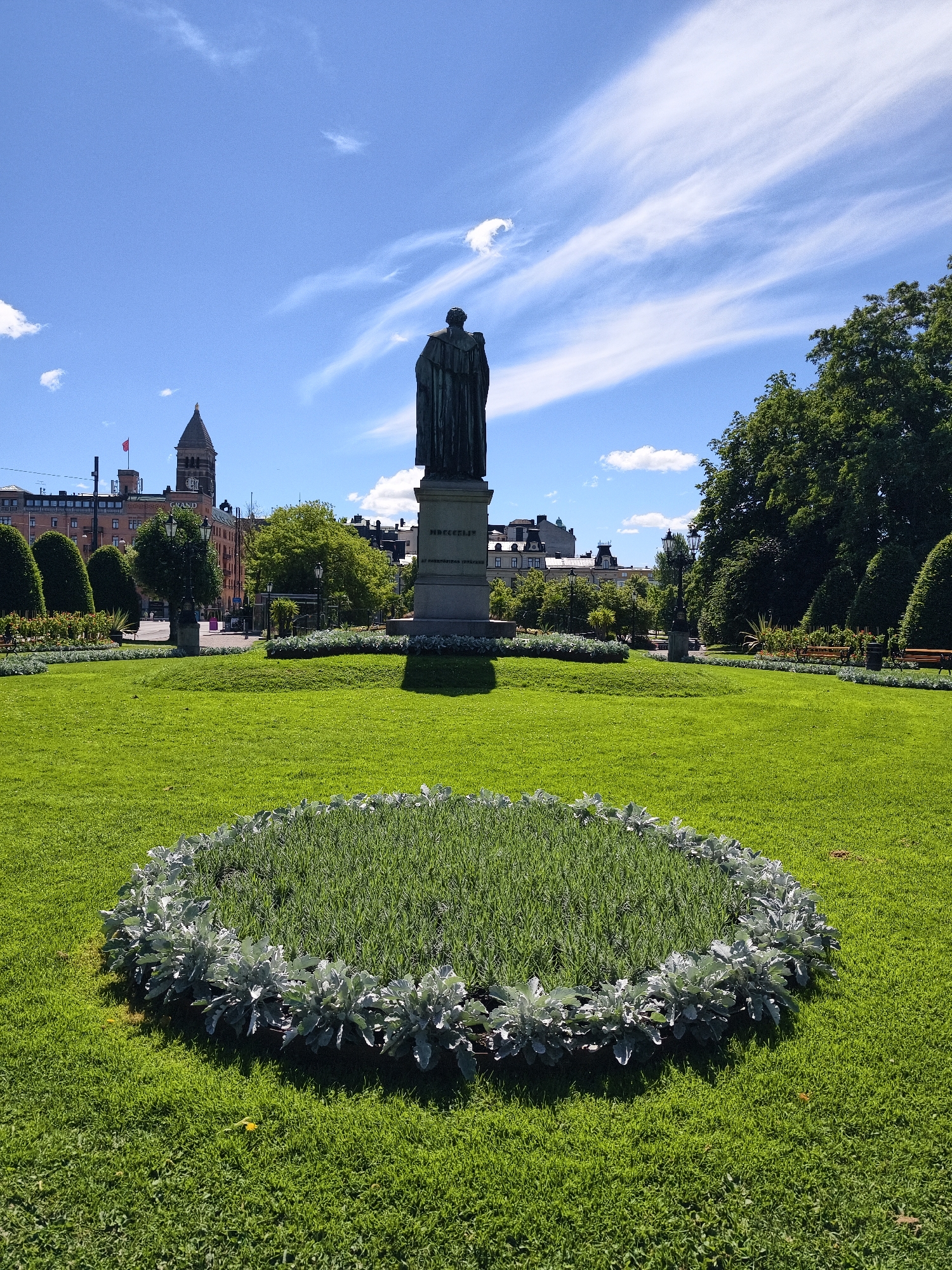 The image depicts a sunny day in a city park, featuring a prominent statue of a robed figure on a pedestal. The statue faces away from the camera, towards the background. In the foreground, a circular flowerbed with pale, silvery-green foliage surrounds a patch of grass. The park is well-maintained with neatly trimmed green lawns and manicured bushes. In the background, several buildings of varying architectural styles are visible, including a tower with a clock and a red flag. Lush green trees frame the scene on the right and in the middle ground. The sky is a clear blue with wispy white clouds. This scene is located in Norrköping, Sweden, specifically in the main square, Gamla Torget, which features the statue of Carl Gustaf Borgström.