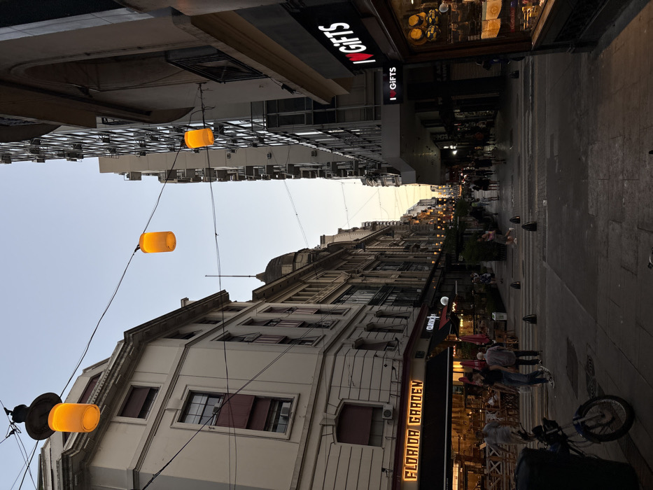 An upside-down view of a bustling street. Buildings line both sides, adorned with glowing cylindrical lights hanging above. A restaurant named "Florida Garden" is visible on the right. Pedestrians stroll along the pavement. A motorbike is parked on the sidewalk. A shop with a sign reading "I <3 GIFTS" stands out on the upper right. The sky is visible between buildings, suggesting twilight.