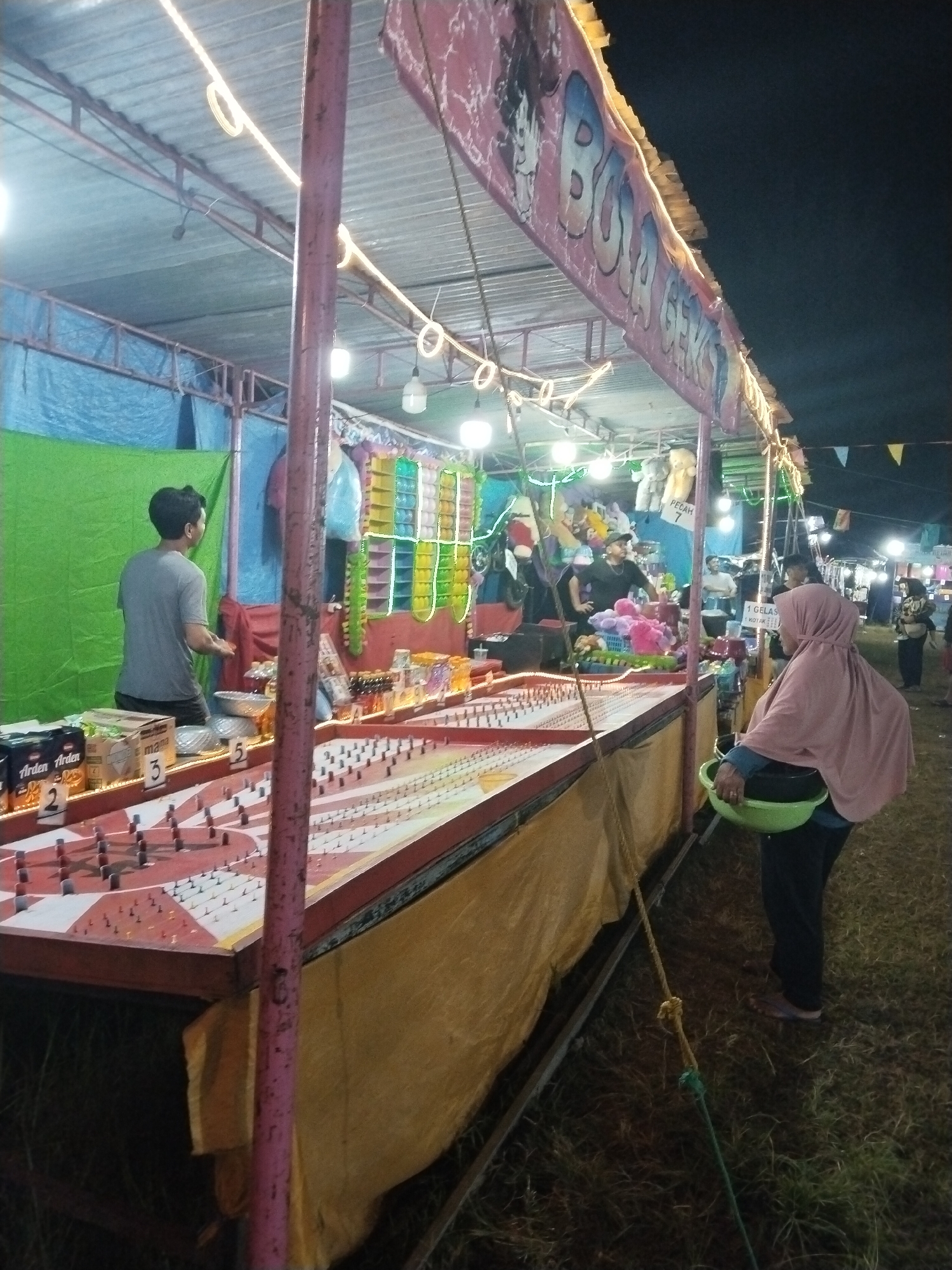 The image depicts a fair or carnival scene at night in Seloromo, Indonesia. A game booth, identified by a sign reading "BOLA GEKSO", is the central focus. The booth features a game where participants aim to hit targets to win prizes, including snacks like "Arden" and "Mama" brand items, and possibly stuffed animals. Two men are operating the booth, one attending to the front and the other handling the back area, presumably managing prizes or stocking. A woman wearing a light-colored hijab stands near the booth, holding a green bowl, possibly containing game pieces or prizes. In the background, other people are visible, suggesting a crowded environment. The booth is illuminated by string lights and bare bulbs, casting light on the game surface, which is patterned with red and white checkerboard designs and dotted with numerous pegs. The ground appears to be grassy.