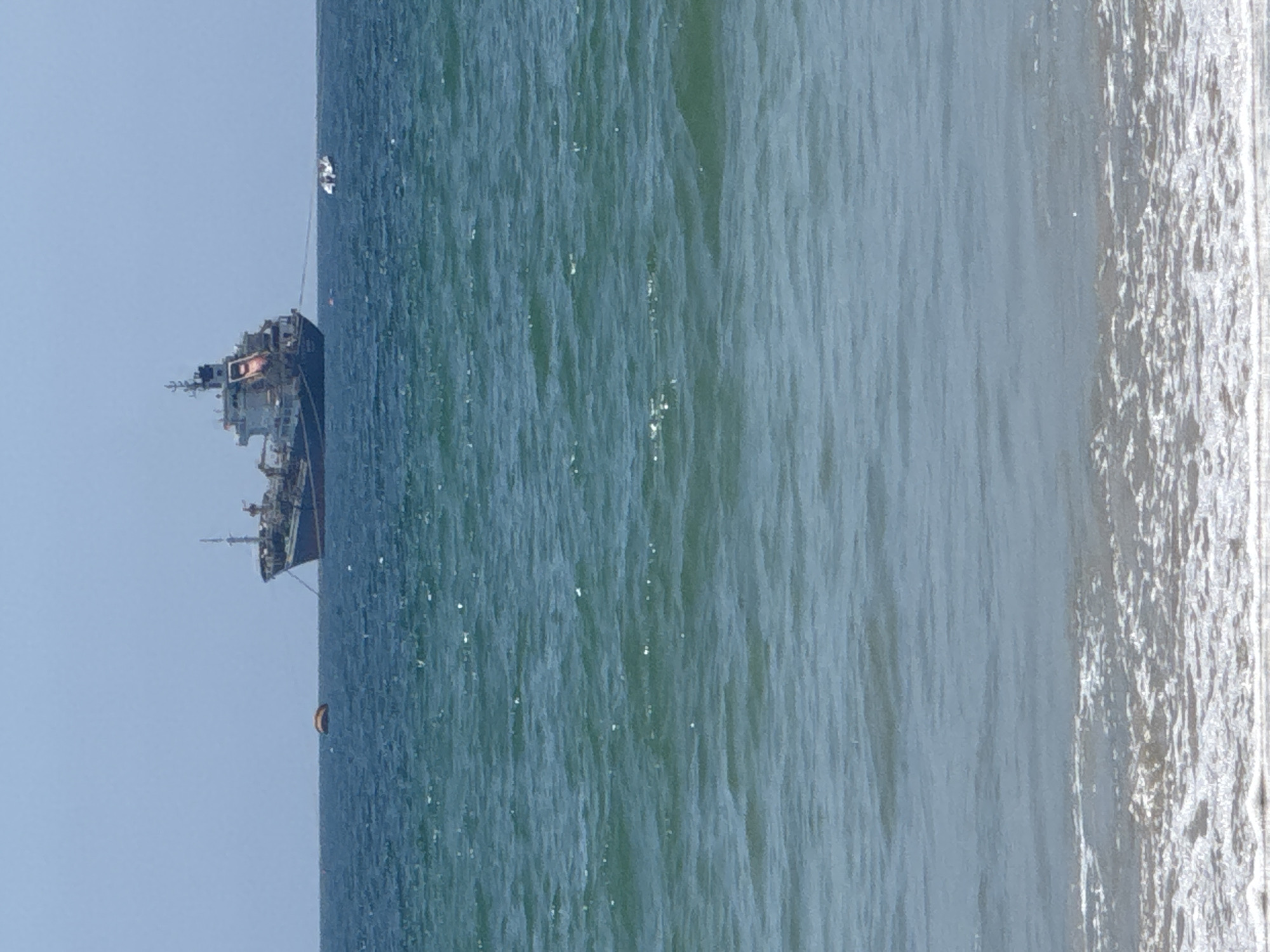 The image, taken in Đà Nẵng, Vietnam, captures a serene coastal scene under a clear, bright sky. The main subject is a large cargo ship or similar vessel, anchored or stationary in the blue-green ocean, positioned towards the left side of the frame. Its superstructure, masts, and portions of its hull are visible, indicating a substantial size.
In the mid-distance behind the ship, a small, white, indistinct object, possibly a buoy or a small tender boat, is visible on the water, appearing to be tethered to the larger vessel by a thin line. Closer to the foreground, in the water to the right of the ship, an orange, crescent-shaped buoyant object, likely a buoy, floats gently.
The ocean water displays subtle ripples and reflections, suggesting calm to moderate sea conditions with sunlight sparkling on the surface. To the right of the image, a sandy shoreline is visible, where gentle, foamy waves break and recede. The sky is a clear, light blue, covering the upper portion of the frame, indicating daytime with good weather. No people are explicitly visible in the scene, and there is no discernible text. The image conveys a sense of tranquility and open space.