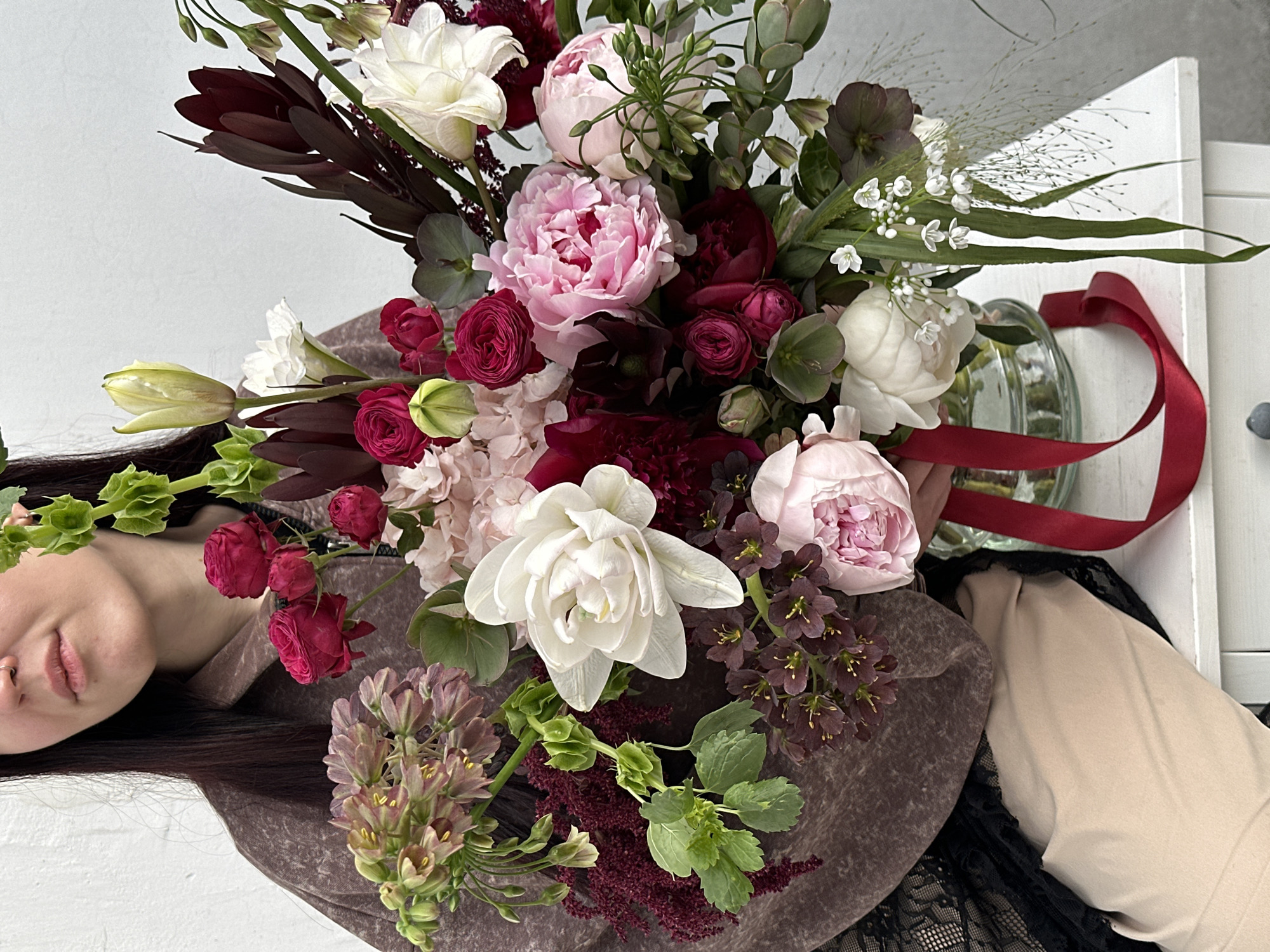 An overhead, slightly tilted shot captures a woman holding a large, opulent floral bouquet against a light background. The woman, partially visible on the left side of the frame, shows her chin, mouth, and nose with a subtle, pleasant expression. She has dark hair and appears to be wearing a textured brown top over a black lace garment.
The dominant subject is the vibrant and diverse bouquet, featuring a rich palette of pink, white, and deep red. Key flowers include large, fluffy pink peonies, dark red garden roses, elegant white double tulips, and clusters of light pink hydrangeas. Darker elements like burgundy protea-like foliage, deep purple hellebores, and burgundy amaranthus add depth and contrast. Green elements such as Bells of Ireland, various broad leaves, and slender grass-like stems with tiny white flowers provide freshness. Clusters of light purple-green bell-shaped flowers are also incorporated.
Behind the bouquet, a clear glass vase or jar is visible, adorned with a bright red ribbon. The background primarily consists of a plain light-colored surface, possibly a wall, with a portion of a white wooden surface appearing on the right. The scene suggests a posed shot, likely showcasing the beauty of the floral arrangement, possibly in celebration or as a gift, in an indoor setting in Moscow, Russia. The lighting suggests it is daytime. No visible text is present in the image.