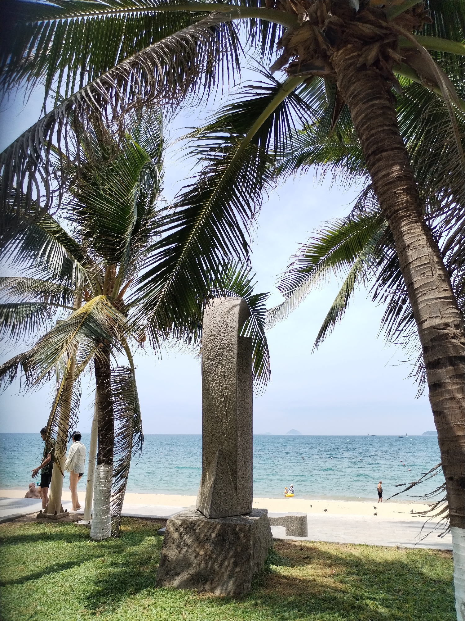 This is a photograph of a beach scene in Nha Trang Ward, Vietnam, taken from a slightly elevated perspective looking towards the ocean. In the foreground, the lush green grass is illuminated by dappled sunlight, casting shadows from the fronds of two prominent palm trees. These palm trees, with their rough, textured trunks and abundant green fronds, frame the view of the beach and the sea.
A stone sculpture, tall and rectangular with a rough, textured surface, stands centrally in the mid-ground, positioned on a stone base. To the left of the sculpture, two individuals are visible near the base of another palm tree. One, wearing a dark t-shirt and shorts, appears to be interacting with another person wearing a light-colored shirt and shorts, who is facing away from the camera.
The sandy beach stretches out beyond a paved walkway, meeting the calm, blue-green ocean. A few people are scattered along the shoreline. One person is visible near the water's edge, appearing to be standing or walking. Further out in the water, another person can be seen, possibly swimming or engaged in a water activity. A yellow inflatable raft is also visible in the shallow water. Several small birds are scattered on the sandy area. The horizon is marked by the meeting of the sea and a hazy sky, with distant islands or landmasses visible on the horizon. The overall atmosphere is peaceful and suggests a sunny day at a tropical beach.