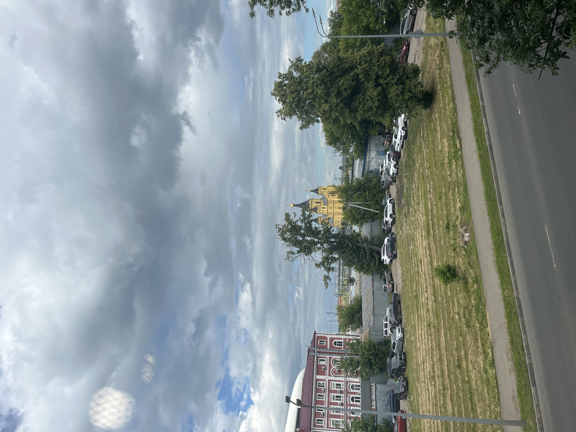 The image captures a street view in Nizhny Novgorod, Russia, under a cloudy sky. In the foreground, a gray asphalt road runs horizontally across the bottom of the frame, with a sidewalk and a strip of dry, mown grass next to it. A street light stands tall on the left side of the road.
Further back, a parking lot is visible, filled with numerous cars. Beyond the parking lot, a distinctive yellow church with multiple domes stands out against the muted sky. To the left of the church, a large, leafy tree is prominent. On the far left of the image, a red building with white trim and arched windows is situated. The overall scene depicts an urban environment with a mix of residential or administrative buildings, a religious structure, and parked vehicles, under a somewhat overcast sky.