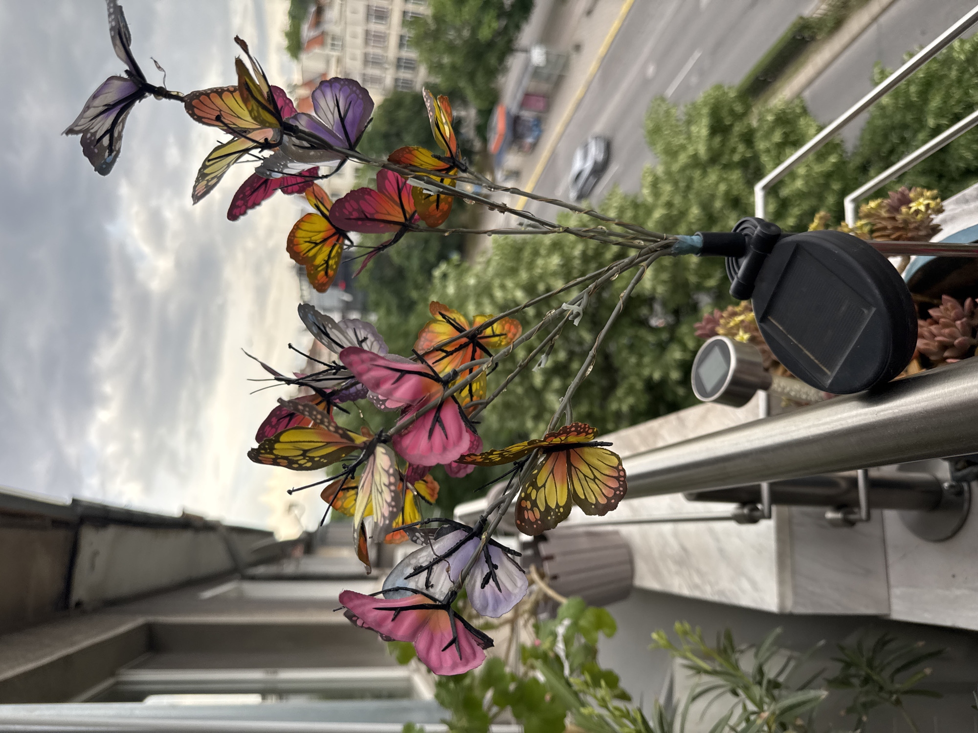 The image shows a close-up of a decorative butterfly light string attached to a balcony railing. The butterflies are brightly colored in shades of pink, yellow, orange, and purple. The light string is powered by a solar panel. The background shows a city scene, including buildings, trees, and a road with cars. Though the city is identified as Plovdiv, Bulgaria, there is no visible indication that this is actually the case.