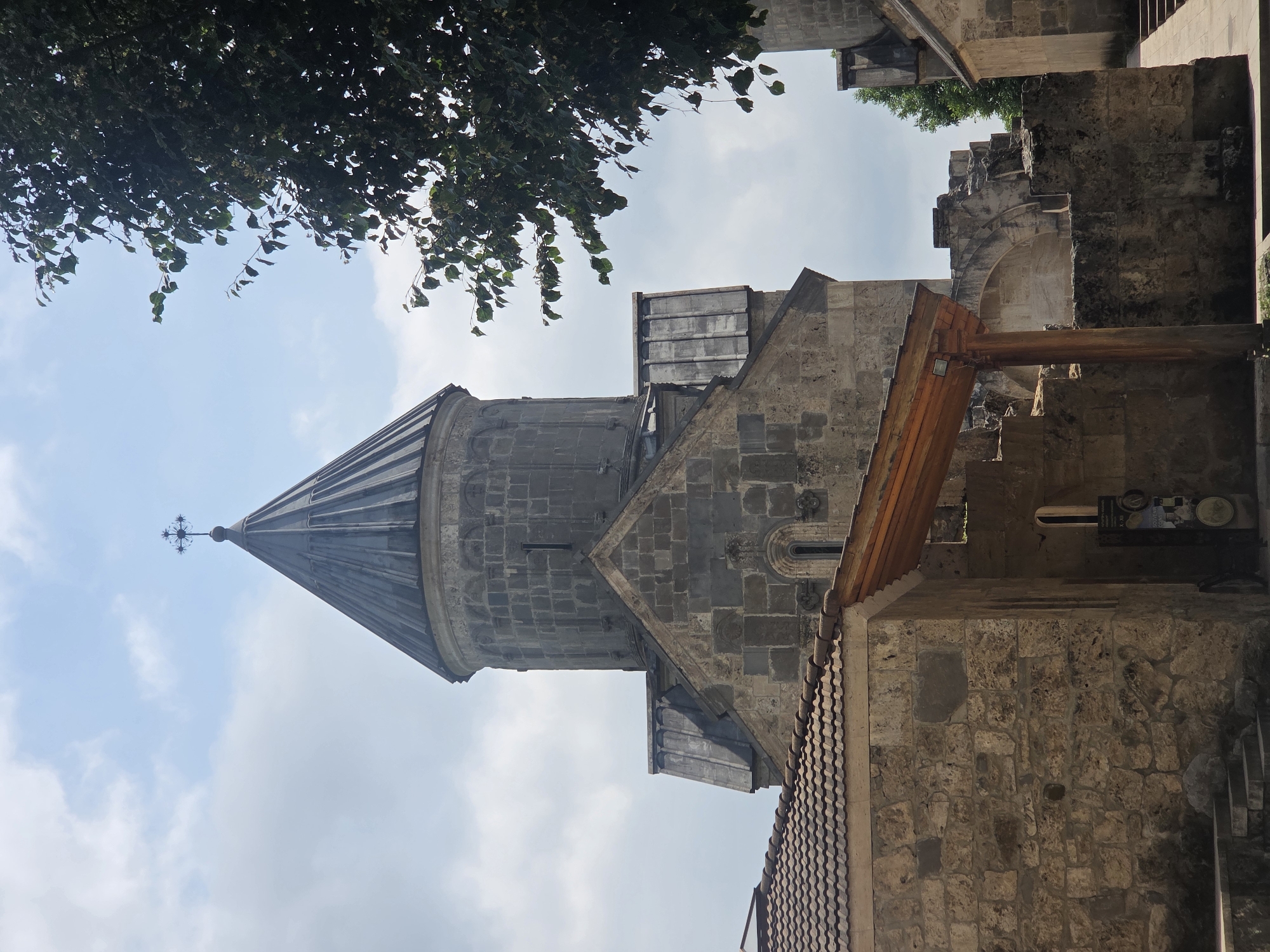 The image shows a stone church with a conical roof topped with a cross. The church appears to be ancient, with weathered stone blocks forming its structure. Part of a tiled roof of a smaller building is visible in the foreground. A tree with dark green leaves is on the right side of the image, partially obscuring the sky. The sky is partly cloudy. The overall scene suggests a historic religious site. Based on the provided information, this is the church in the village of Teghut, Armenia. There are no people visible in the image.