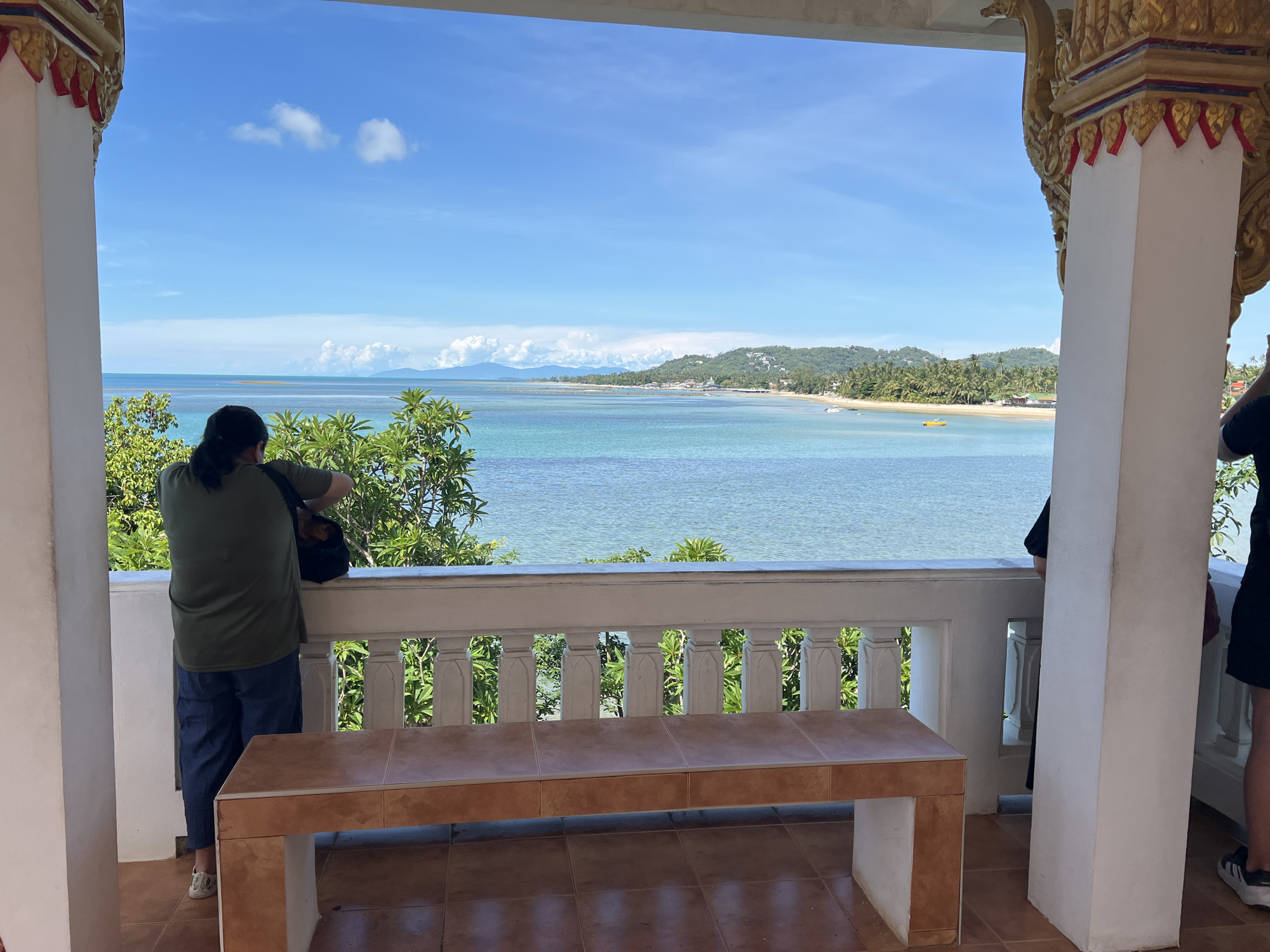 The image depicts a serene view of the ocean and coastline from a covered balcony, likely in Baan Hua Thanon, Thailand. In the foreground, a person wearing a green t-shirt and blue pants stands with their back to the camera, looking out at the water. To the right, another person's legs are visible, wearing black shorts and white-soled athletic shoes. A long, low bench with a tiled surface sits in the center of the balcony. The balcony itself is characterized by a white railing with decorative balusters and ornate golden architectural details on the supporting columns. Beyond the balcony, the turquoise ocean stretches to the horizon, meeting a pale blue sky dotted with a few white clouds. The distant coastline features lush green hills covered in palm trees and scattered buildings. A yellow boat is visible in the water. The overall atmosphere is tranquil and summery.