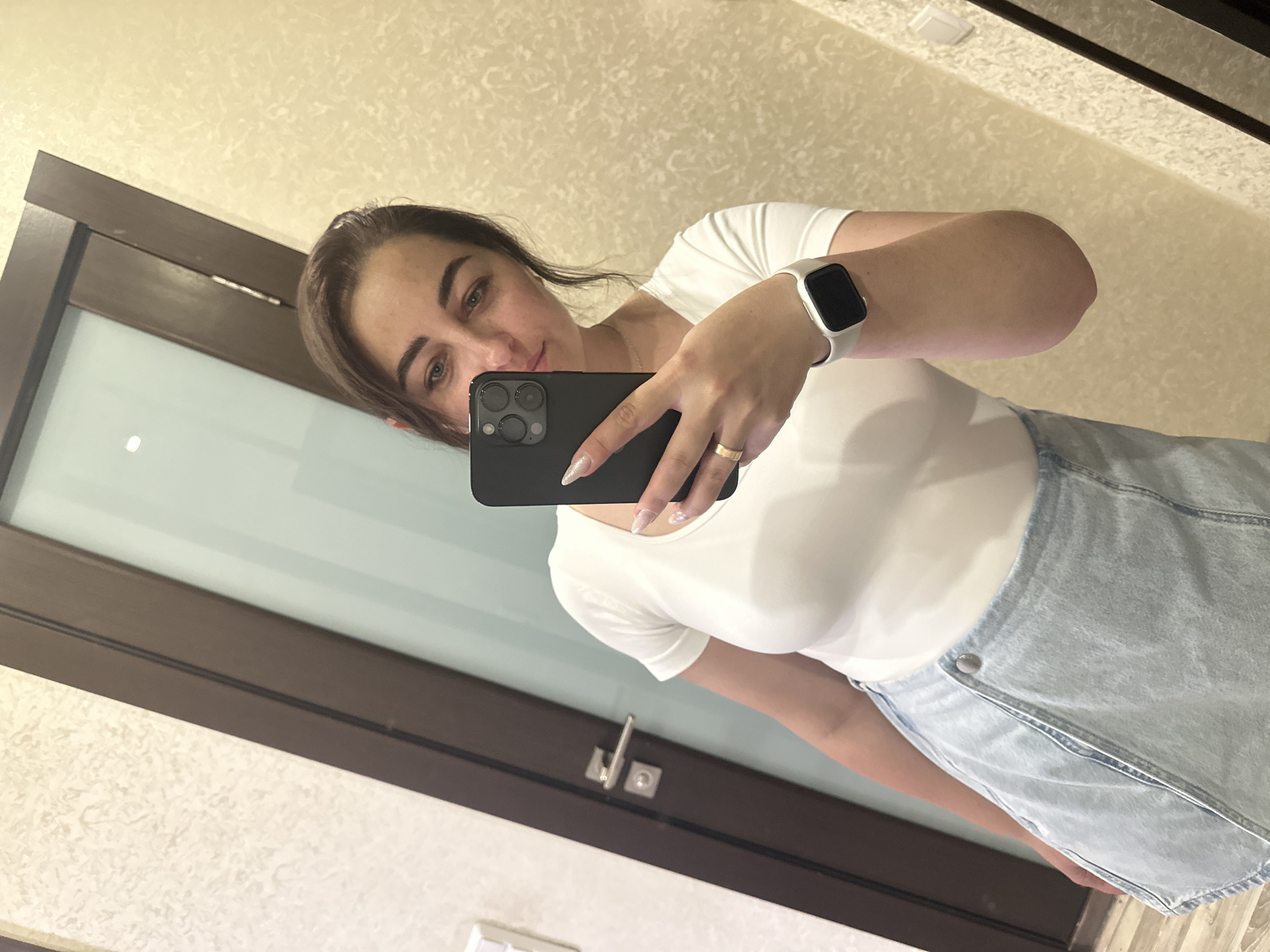 A woman stands in a dimly lit room, taking a selfie in a mirror. She is wearing a white t-shirt and a light blue denim skirt. On her left wrist, she wears a white smartwatch. Her right hand holds a black smartphone, angled to capture her reflection. The background shows a door with a silver handle and a textured wall. Based on the available data, the image was captured in Shchyolkovo, Russia.