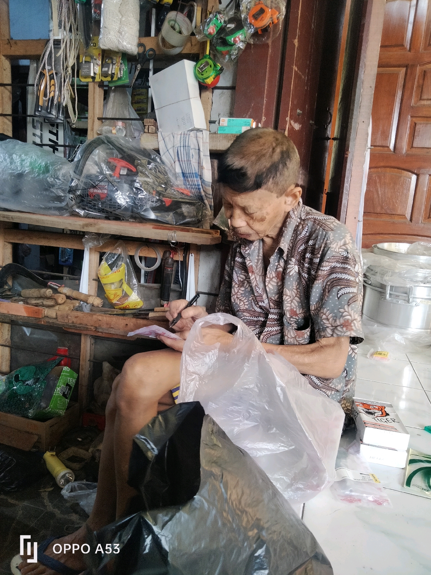In the city of Ngulanwetan, Indonesia, an elderly man with a patterned shirt sits on the floor in front of shelves filled with various items, including tools, rope, and packaged goods. He is holding a pen and appears to be writing on a piece of paper, possibly a receipt or a list, while a large plastic bag sits in front of him. The environment suggests a cluttered storage area or a small shop.