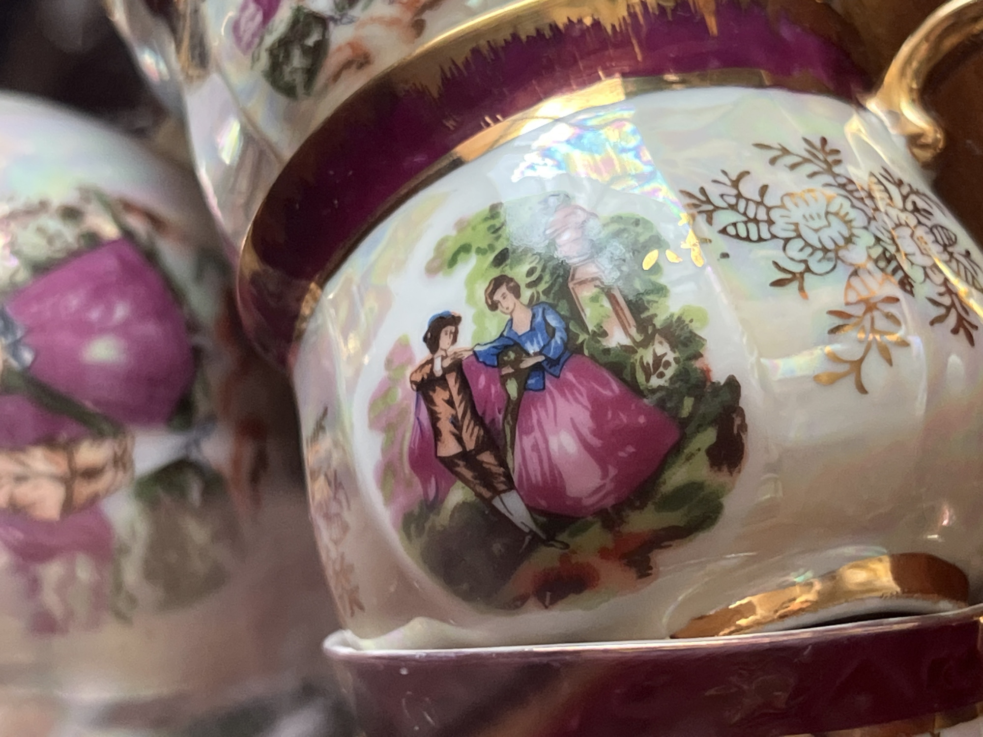 The image shows a close-up of a stack of decorated teacups. The cups are white with iridescent sheen and feature painted scenes of figures in a garden or park setting. One scene depicts a man and woman standing next to a lamp post, with the woman wearing a pink dress and blue shawl, and the man in a brown suit. The cups are trimmed with gold and maroon detailing. The location is not identifiable from the image.