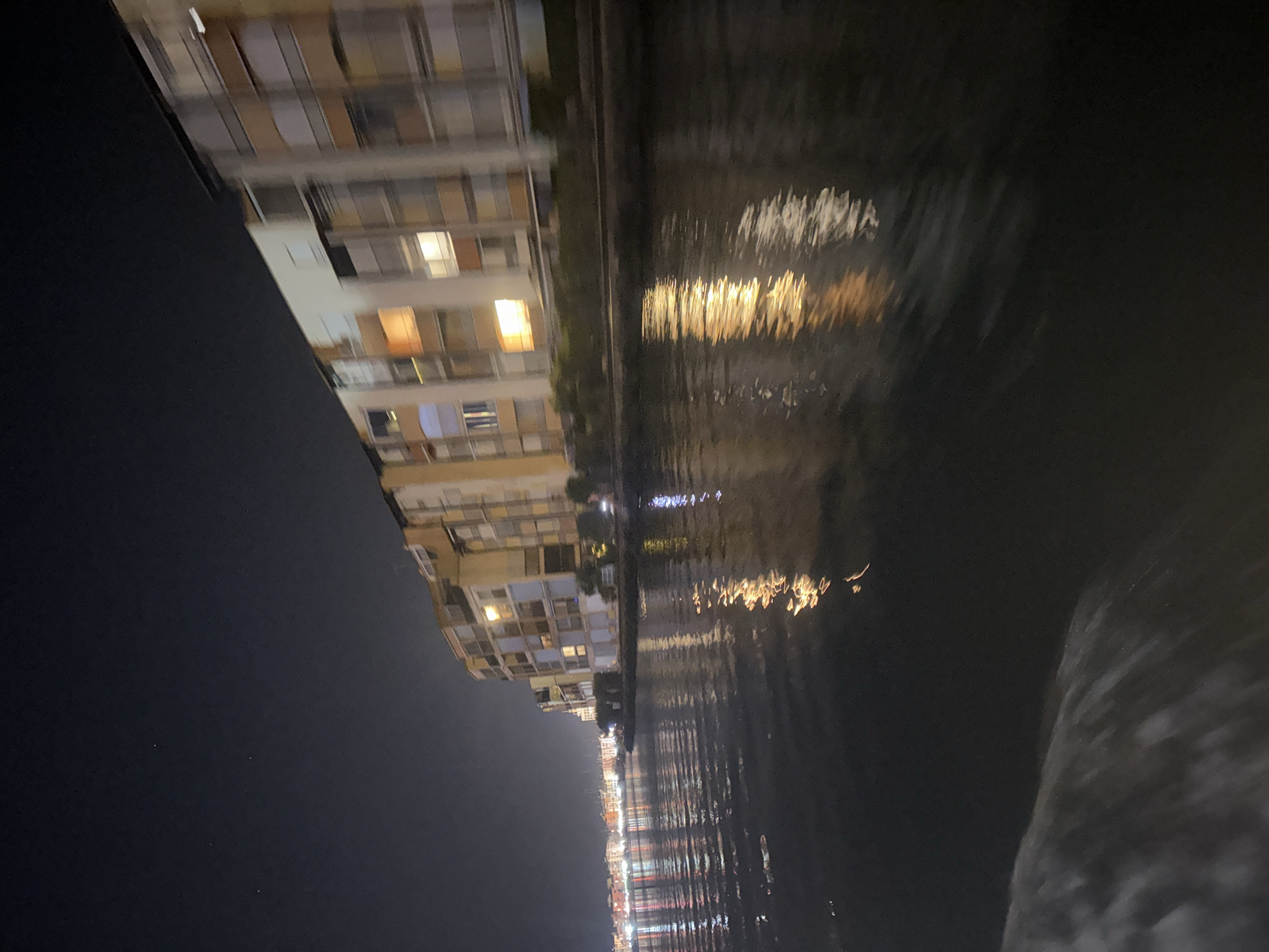 This is a nighttime, medium-shot, slightly blurry photograph taken from a body of water, likely a canal or river, looking towards a waterfront of buildings and the sky. The water in the foreground is dark and reflects the lights from the buildings, creating shimmering streaks of orange, yellow, and white. A white wake is visible in the lower left corner, indicating movement.
The buildings are a mix of multi-story structures with balconies and windows, illuminated from within. The architecture appears modern, with light-colored facades and various window designs, some with closed shutters. The buildings stretch along the waterfront, receding into the distance on the left where more lights are visible, suggesting a populated area. The sky above is dark with a few faint stars visible.
The scene is located in Agde, France, likely along the Hérault River or the Canal du Midi, given the presence of waterfront buildings and the reflective water. The overall impression is one of a tranquil, illuminated waterfront at night. There are no people clearly visible in the image.