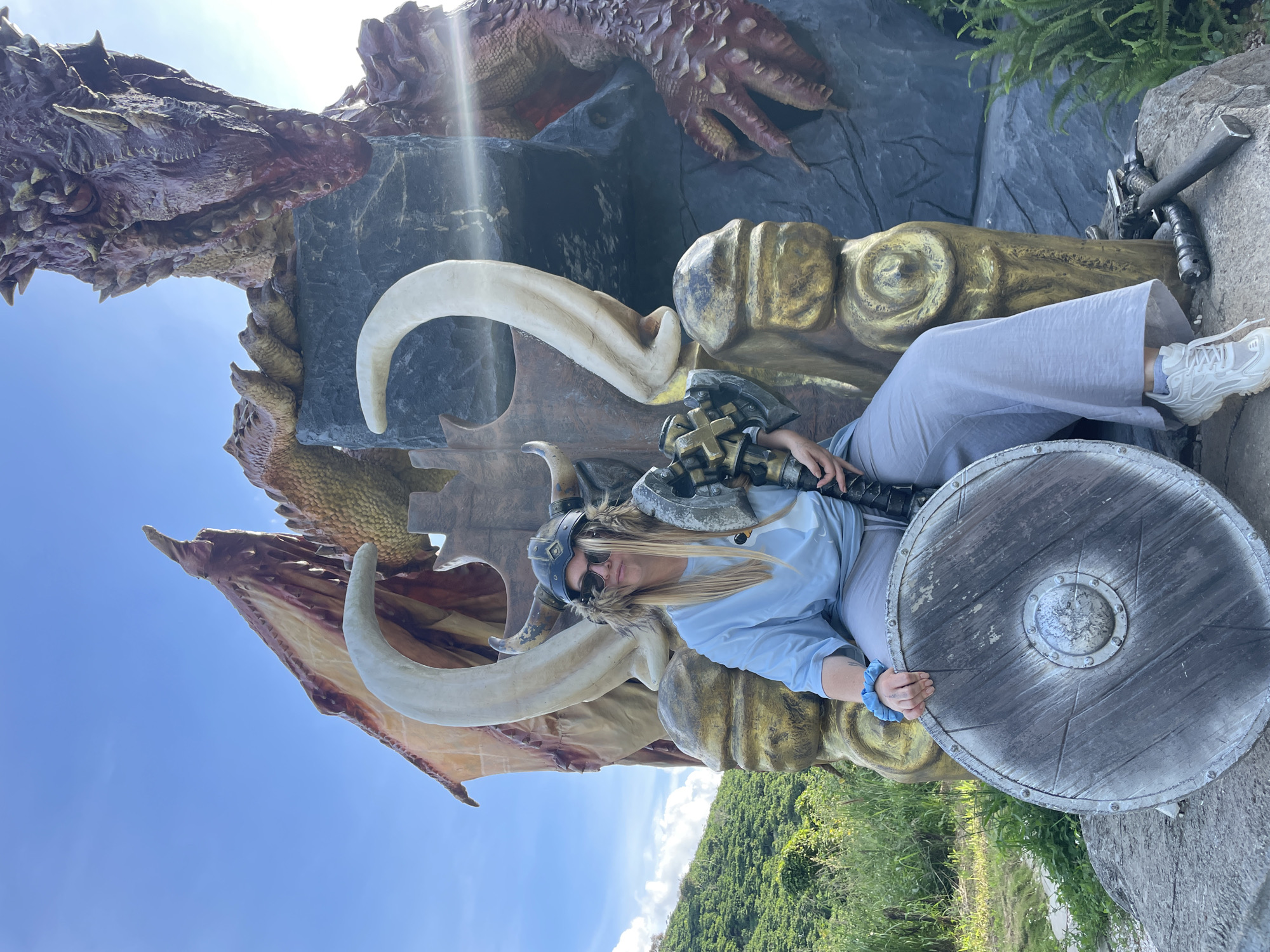 A person sits on a throne in front of a dragon statue. The person is wearing a Viking helmet with fur trim, sunglasses, a blue t-shirt, and grey pants. They are holding a large axe in their right hand and a round shield on their left. The throne is made of gold-colored material and appears to be carved to resemble tusks and possibly a rough, rocky texture. The dragon statue is large and reddish-brown with prominent scales and wings. Behind the throne, a rocky structure is visible, with a portion of the dragon's body and head appearing to emerge from it. The background shows a bright blue sky with some scattered clouds, lush green hills covered in vegetation, and some smaller green plants in the foreground. The overall scene suggests an outdoor attraction or themed environment, possibly a tourist spot. No specific location within Thôn 1, Vietnam is identifiable from the image alone.