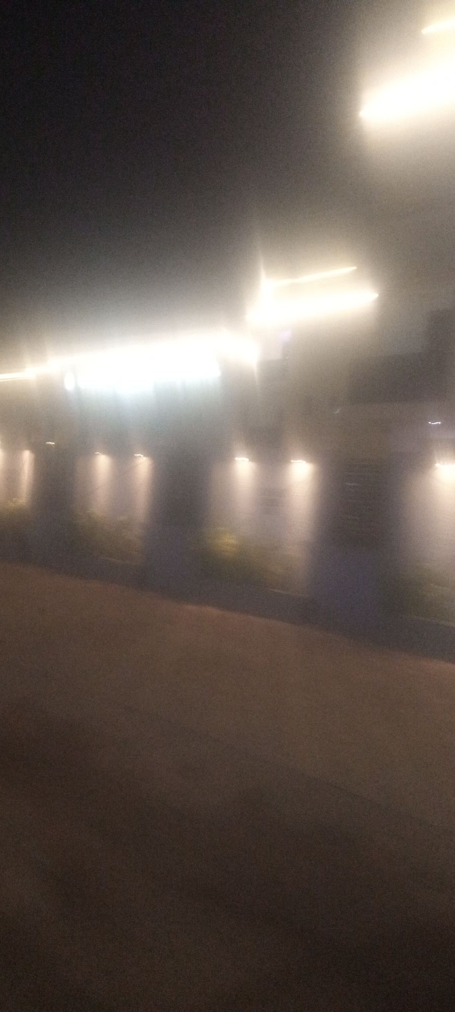 The image appears to be taken at night in Narayi, Nigeria, though specific location details within the city are unclear. There are lights illuminating what appears to be the exterior of a building or a series of structures along a road. The image is blurry, suggesting movement during the photo capture. The ground is paved, with what seems to be a curb or landscaping bordering the building.