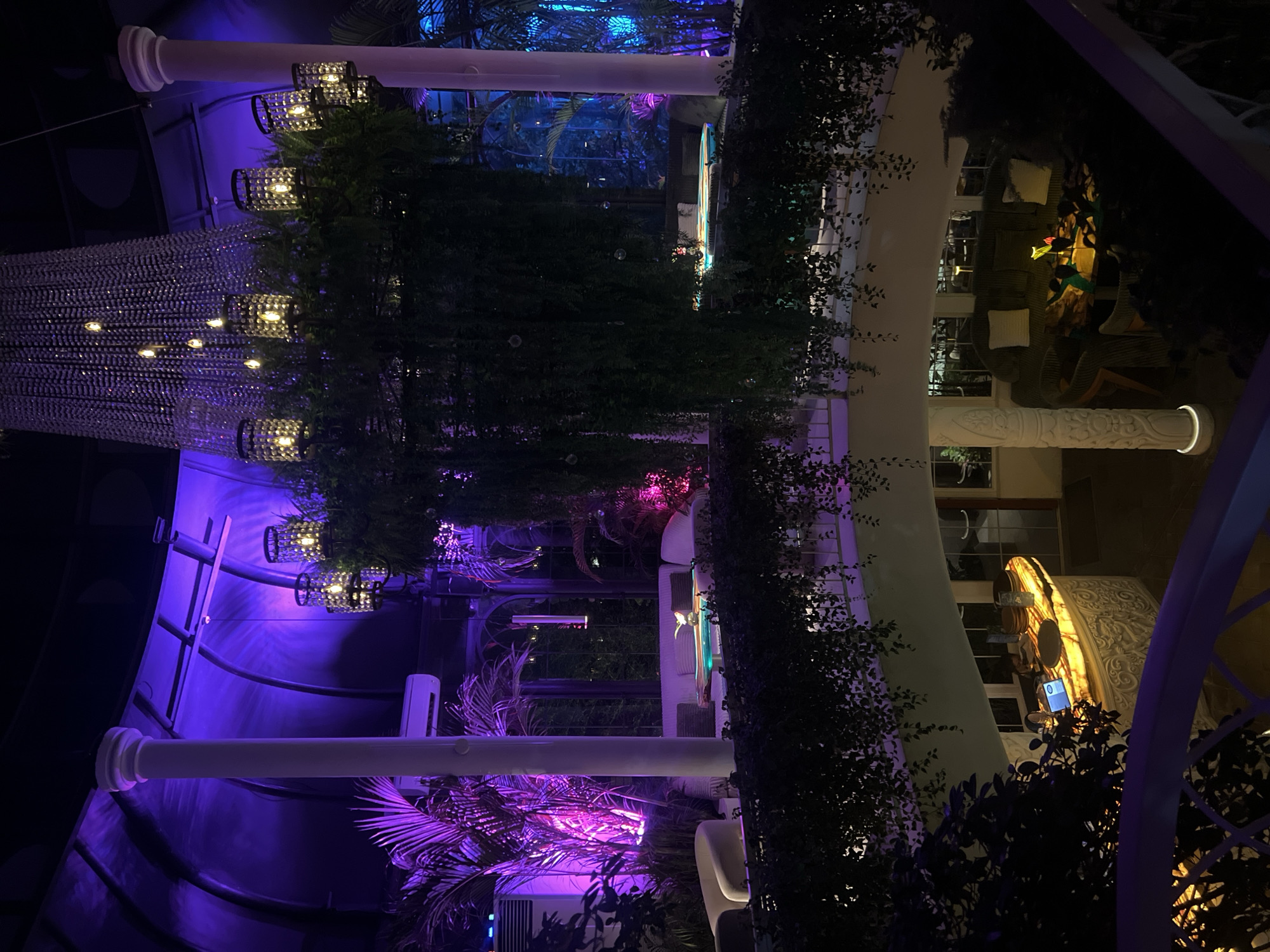 The image depicts the interior of a building, likely a restaurant or lounge, located in North Kuta, Indonesia. The scene is lit with a combination of warm and cool lighting, predominantly purple and yellow hues. Architectural elements include white columns, arched frameworks, and ornate wall carvings. Lush greenery, including hanging plants and palm trees, is integrated into the design. A large chandelier hangs from the ceiling. Furniture, such as chairs and tables, is visible, but no people are clearly visible in the frame. The overall impression is one of an upscale, aesthetically designed space.