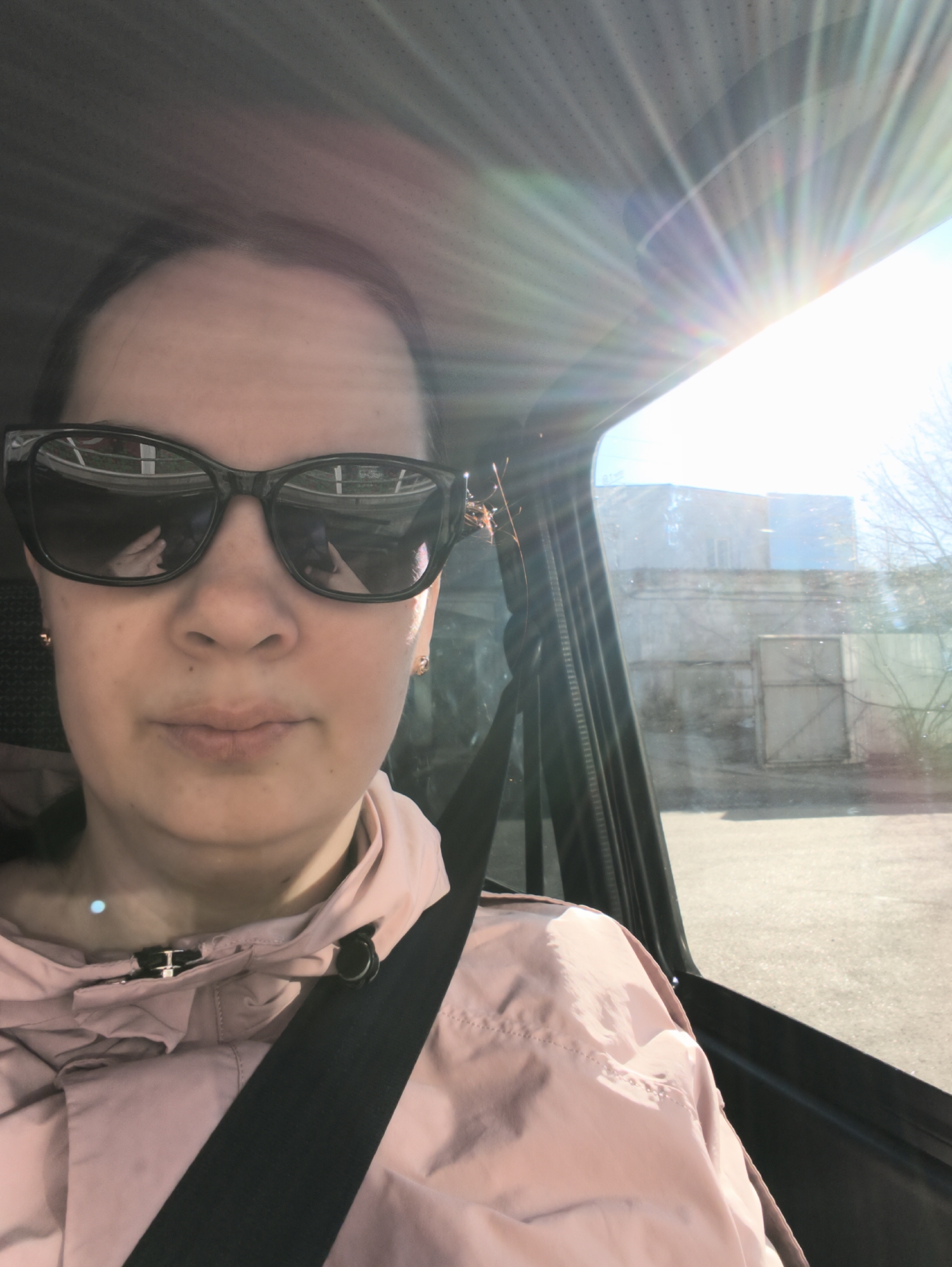 A woman wearing sunglasses and a pink jacket with a seatbelt is in a car. The sun is shining brightly through the car window, behind which is a building and a road. The location is Unknown, Unknown.