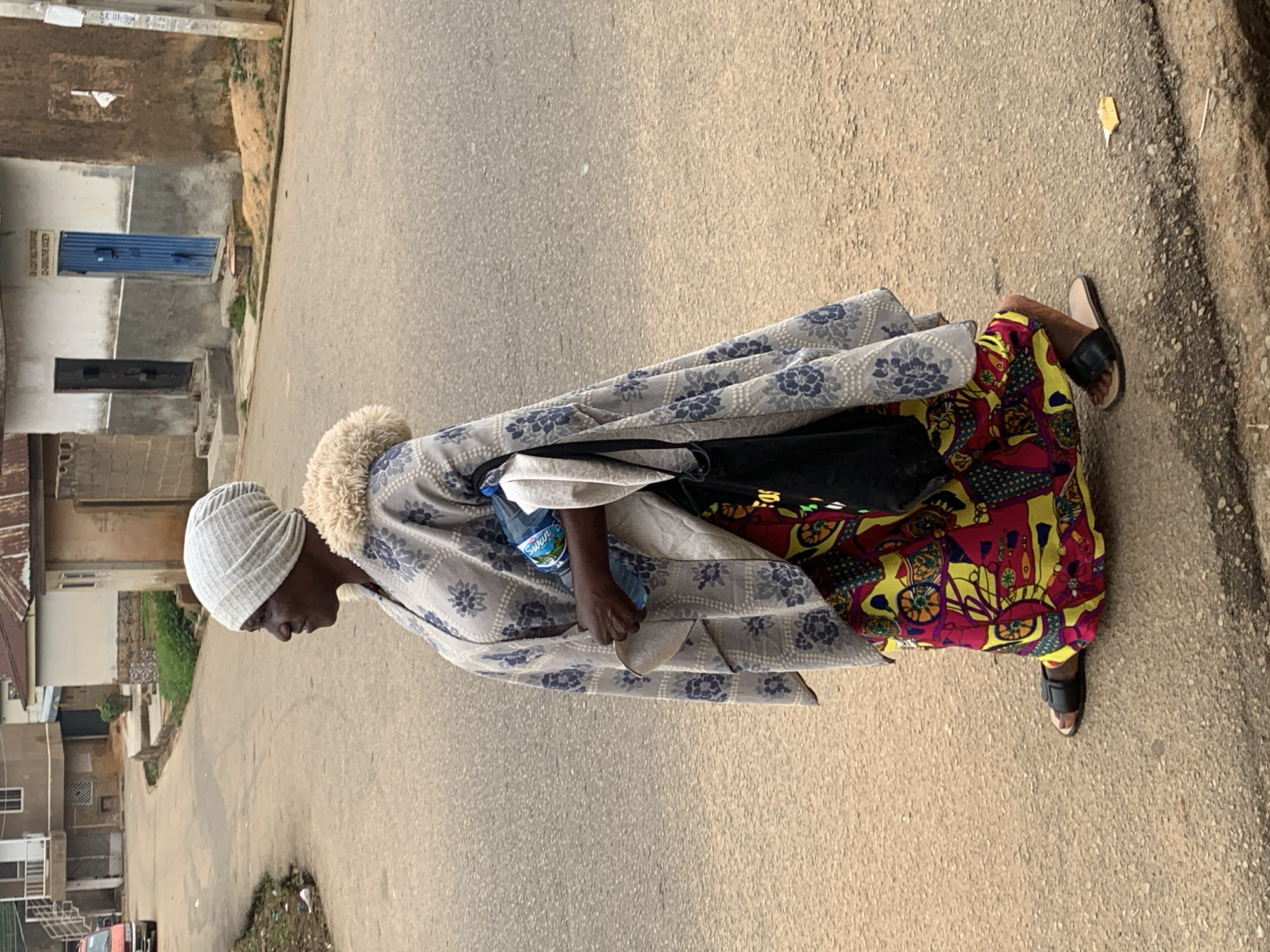 A full-body, side-view, medium-distance shot captures a woman walking on a paved street in Jos, Nigeria. She is facing left, wearing a light gray knitted headscarf, a light gray, patterned shawl with a fluffy cream-colored collar, and a vibrant, multi-colored long skirt with a yellow, red, and blue pattern. She is holding a blue plastic water bottle in her right hand and has a black bag slung over her left shoulder. She is wearing dark-colored open-toed sandals. The street is asphalt, with a visible curb. Buildings line the street, including a two-story building with a red vehicle parked to its left and another single-story building with a blue door and a sign above it. The environment appears to be a residential or commercial area of the city.