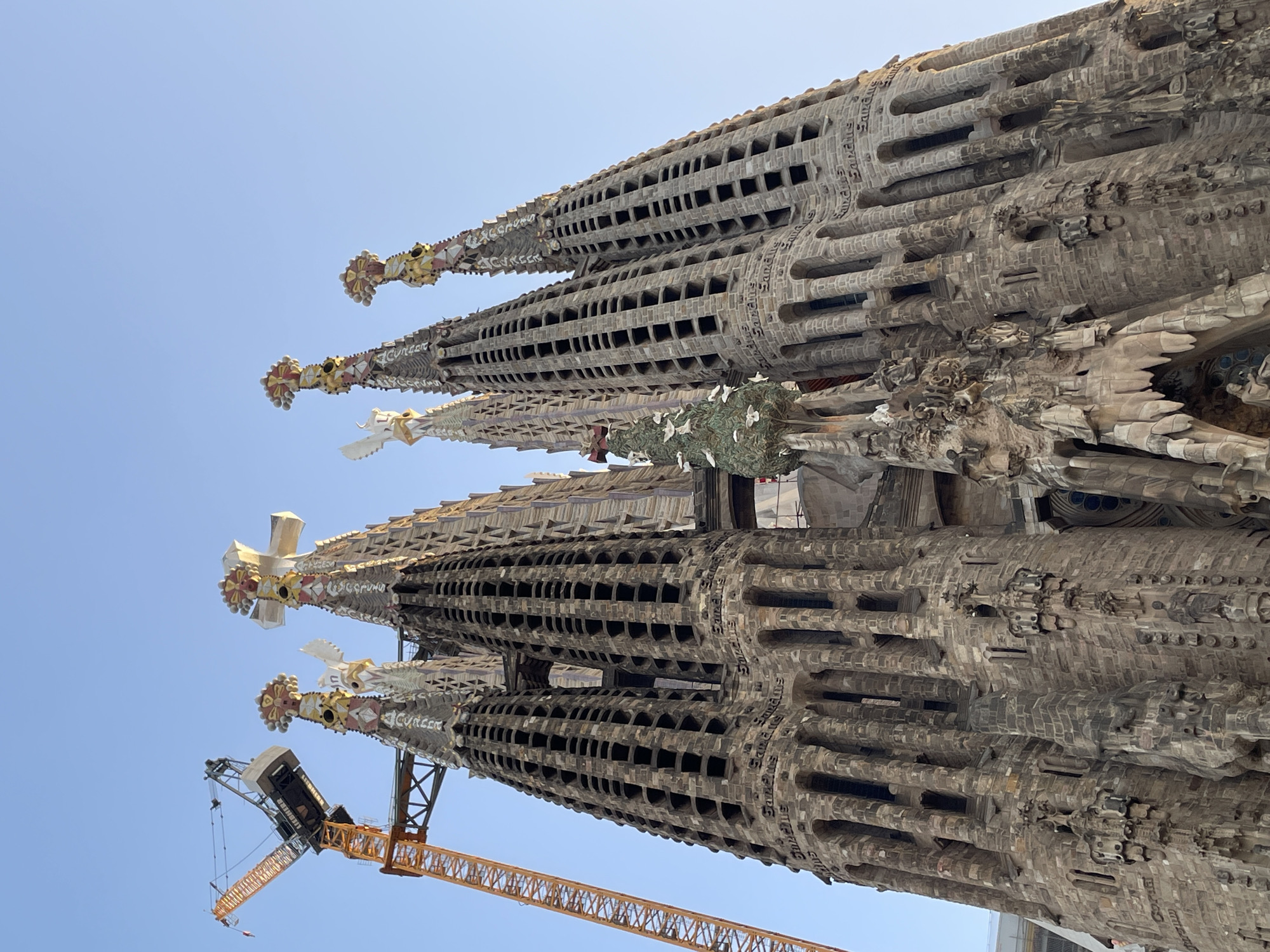 The image shows a low-angle view of the Sagrada Familia basilica in Barcelona, Spain, under a clear blue sky. The basilica's distinctive spires, adorned with intricate carvings and religious symbols, dominate the frame. A large yellow construction crane is visible to the left, indicating ongoing construction or renovation work on the iconic structure. The detailed stonework of the basilica, with its numerous columns, arches, and sculptures, is clearly visible. The overall scene captures the grandeur and ongoing development of this famous architectural landmark.