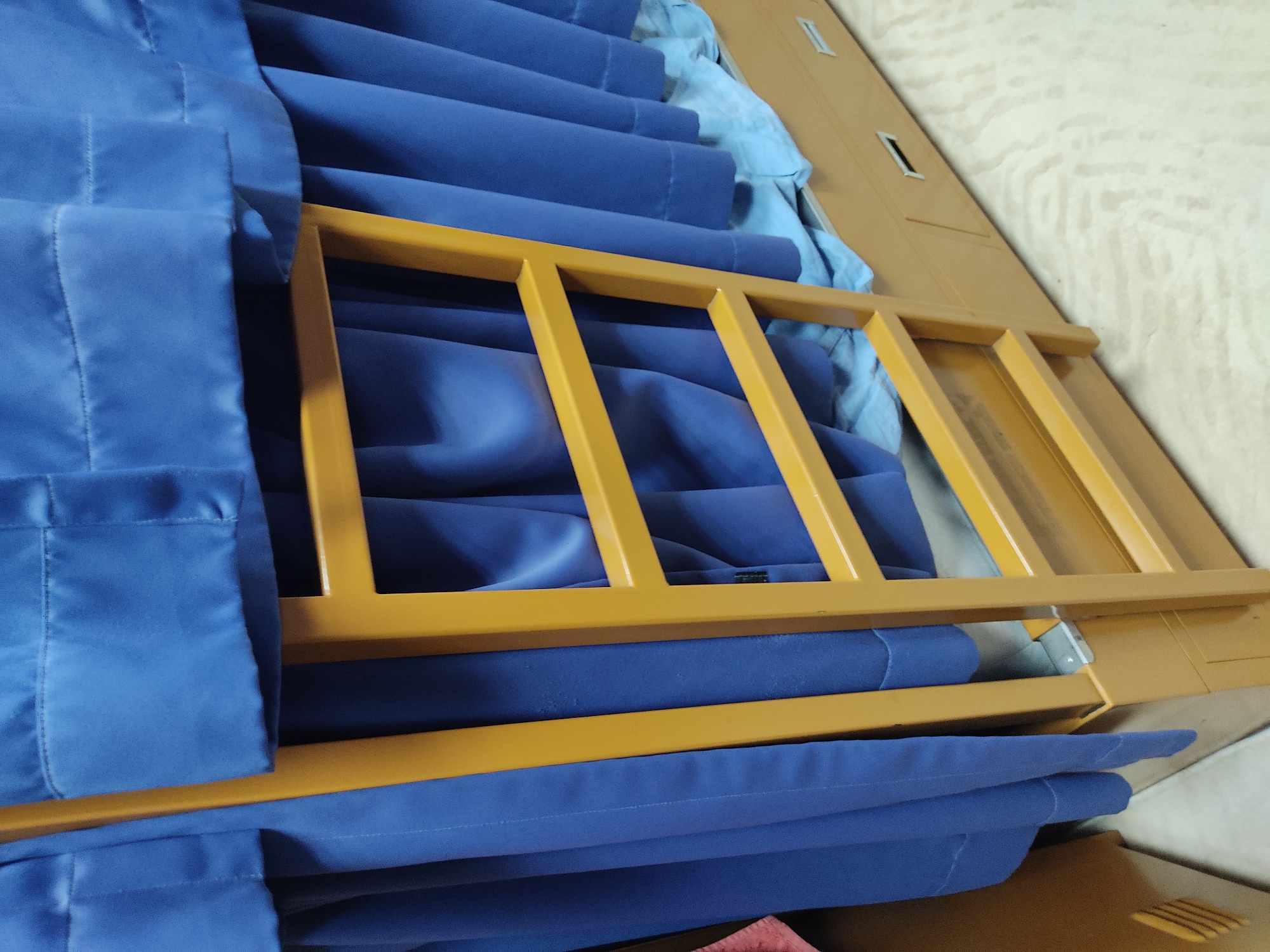 A close-up, angled shot shows stacked blue fabric, possibly blankets or curtains, arranged in neat rows. A yellow metal railing, appearing to be part of a bunk bed or a similar structure, runs horizontally across the frame, partially obscuring the fabric. The fabric is a deep, rich blue with visible stitching and folds. Some of the fabric has a lighter blue lining or is a lighter shade. The railing has a matte, slightly worn appearance. The background is out of focus, showing a textured, light-colored surface, possibly a wall or a floor, with faint wood grain patterns. There are a few small, rectangular indentations on a larger, tan-colored rectangular object in the upper right of the frame, which is also out of focus and appears to be a piece of furniture or packaging. The lighting is soft and appears to be artificial, creating gentle shadows. There is no visible text. The scene suggests an indoor setting, possibly a storage area, a dormitory, or a theatrical space. The stacked fabric and railing imply a utilitarian purpose.