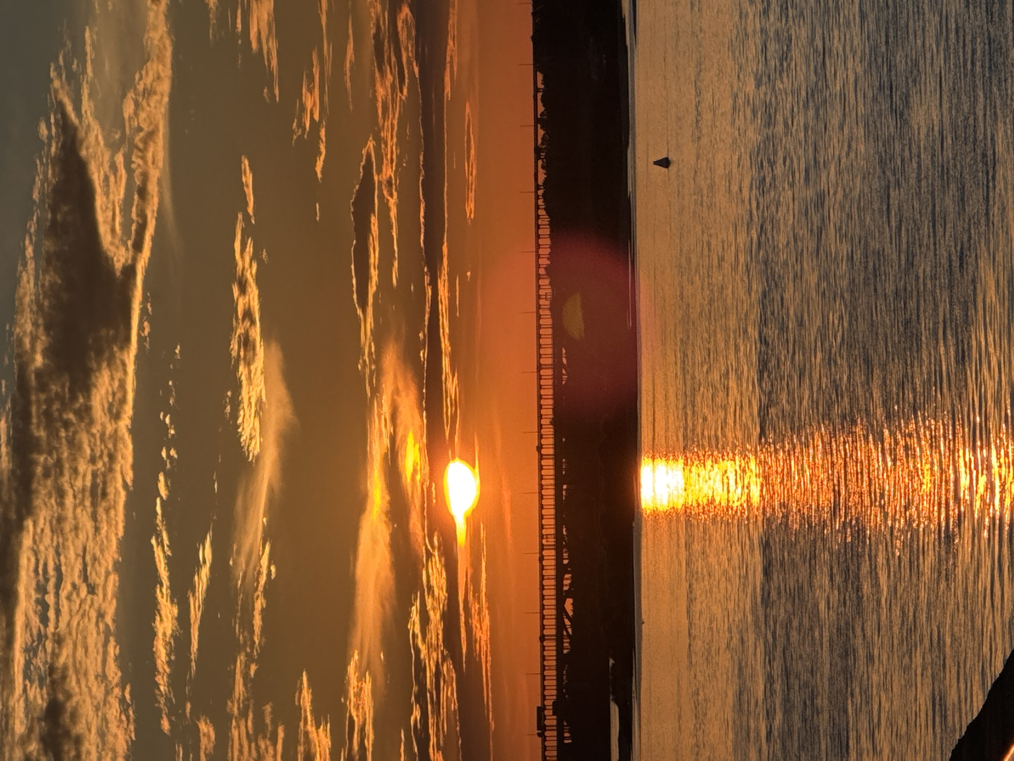 The image depicts a sunset over a body of water in Nizhny Novgorod, Russia. A long bridge with numerous arches stretches across the horizon, silhouetted against the vibrant orange sky. The sun, a bright orb, is partially obscured by clouds, casting a golden reflection on the rippling water. A small buoy floats in the distance. The scene is dominated by the warm hues of the sunset and the dramatic silhouette of the bridge.
