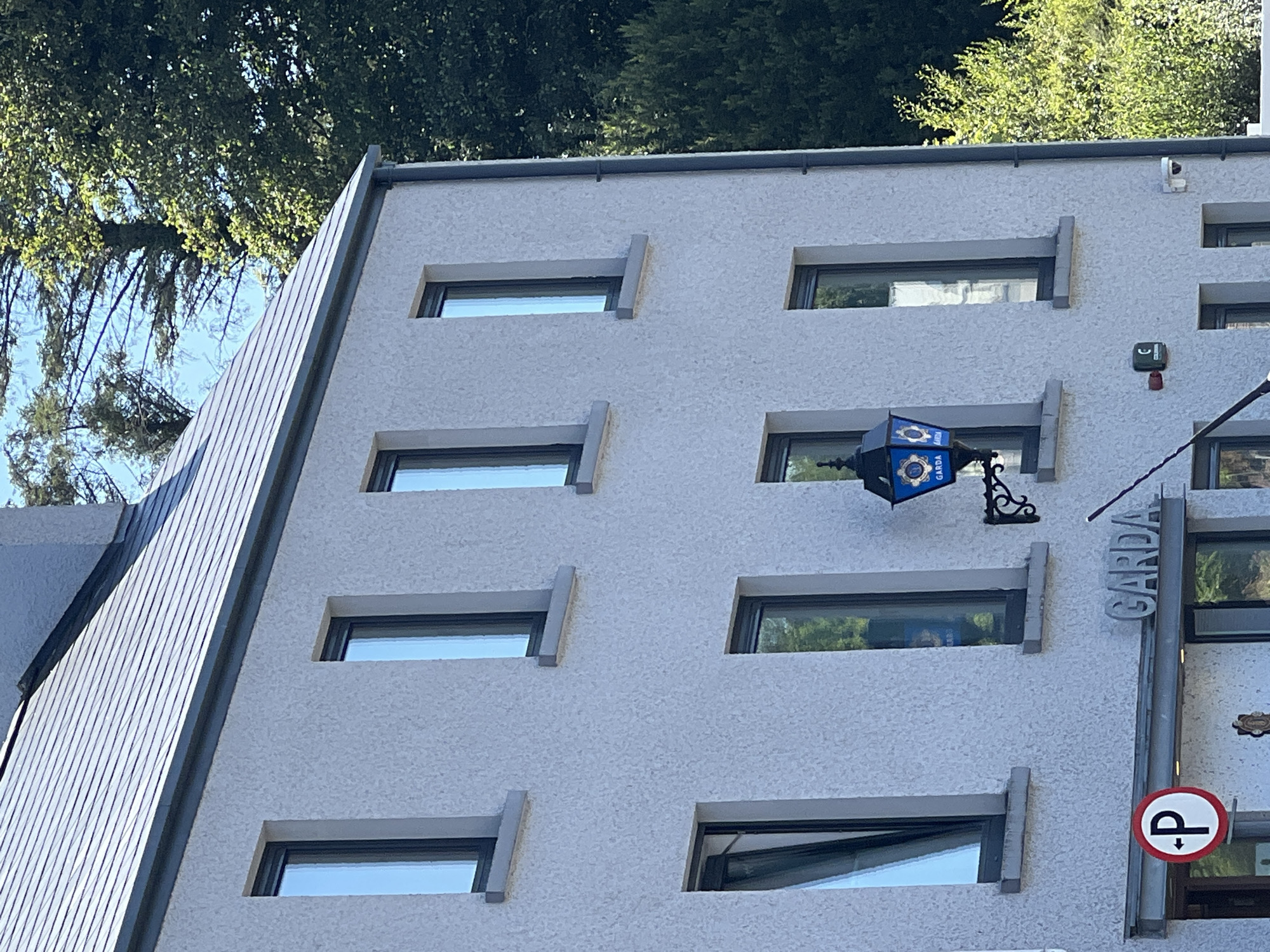 This is a frontal, medium-shot photograph of the exterior of a two-story building, likely a police station, located in the town of Donegal, Ireland. The building is constructed of light-colored stucco and features a dark-tiled roof. Multiple rectangular windows are visible on both stories. A blue and gold lantern with the word "GARDA" is mounted to the wall between two windows on the second story. Below this, on the first story, is a sign with the word "GARDA" in raised silver lettering. To the left of this sign, a red and white circular parking sign with a white "P" is visible. A security camera is mounted on the wall to the right of the "GARDA" sign. Trees and foliage are visible in the background, suggesting an outdoor, daylight setting. There are no people visible in the image.