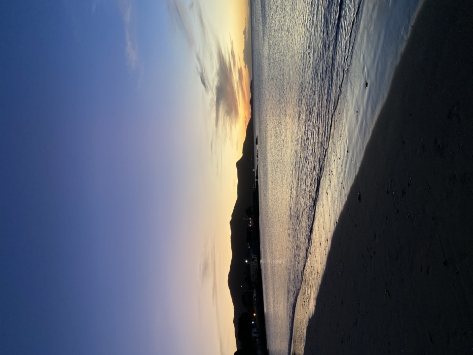 A serene beach scene unfolds during the sunset. Mountains stand silhouetted against the horizon, where the sky transitions from a bright orange glow to a soft blue. Gentle waves reach the shore, reflecting the fading light. The sand in the foreground, cast in shadow, adds depth and contrast to the tranquil vista.