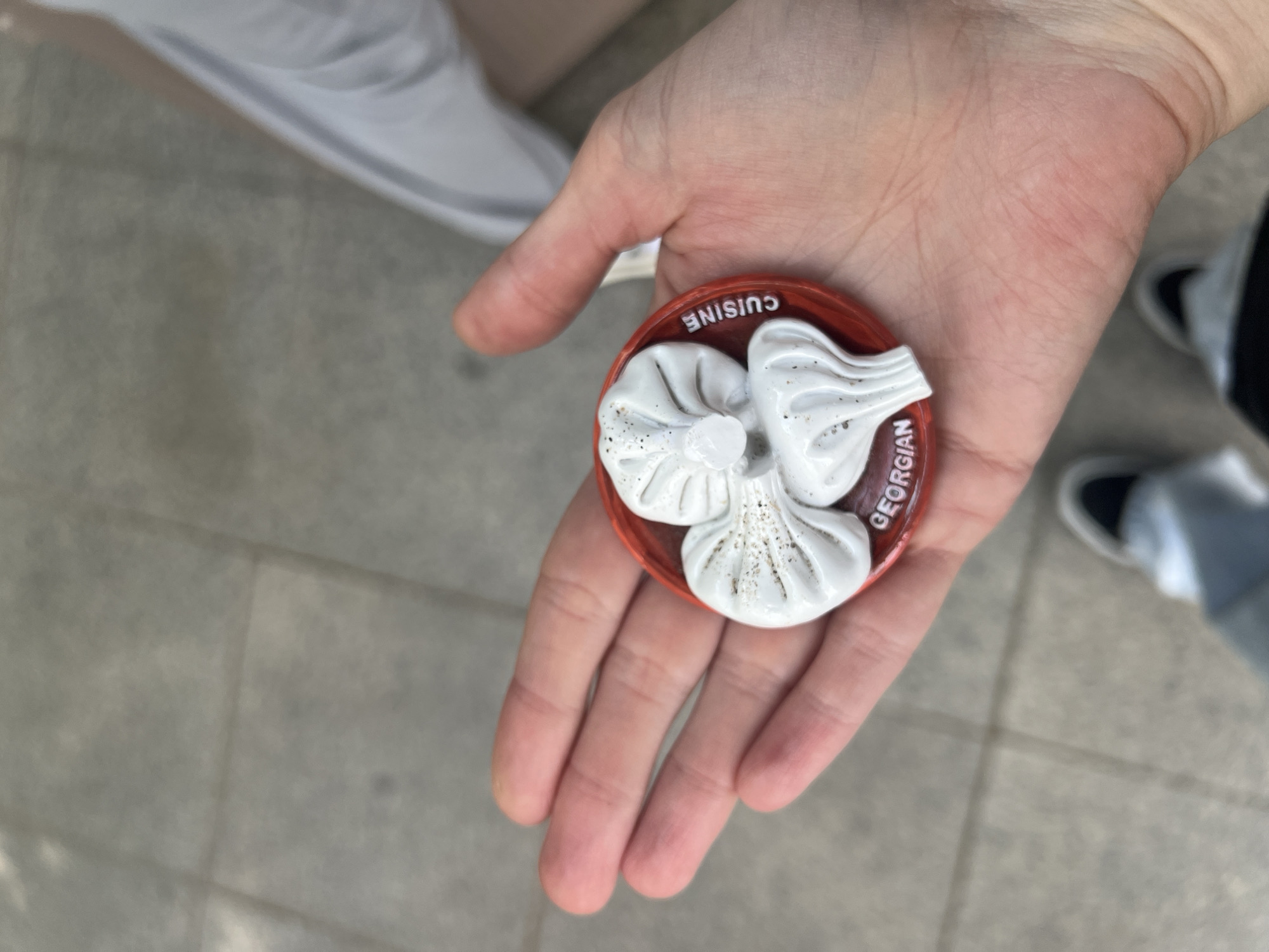 A person holds a Georgian cuisine souvenir in their hand. The souvenir is a round, red object with three white khinkali (Georgian dumplings) molded on top. The words "Georgian Cuisine" are printed on the souvenir. The person is standing on a tiled surface, possibly a sidewalk, and parts of their white sneakers and other shoes are visible in the background. The scene may be in Tbilisi, Georgia, due to the prominent display of Georgian dumplings.