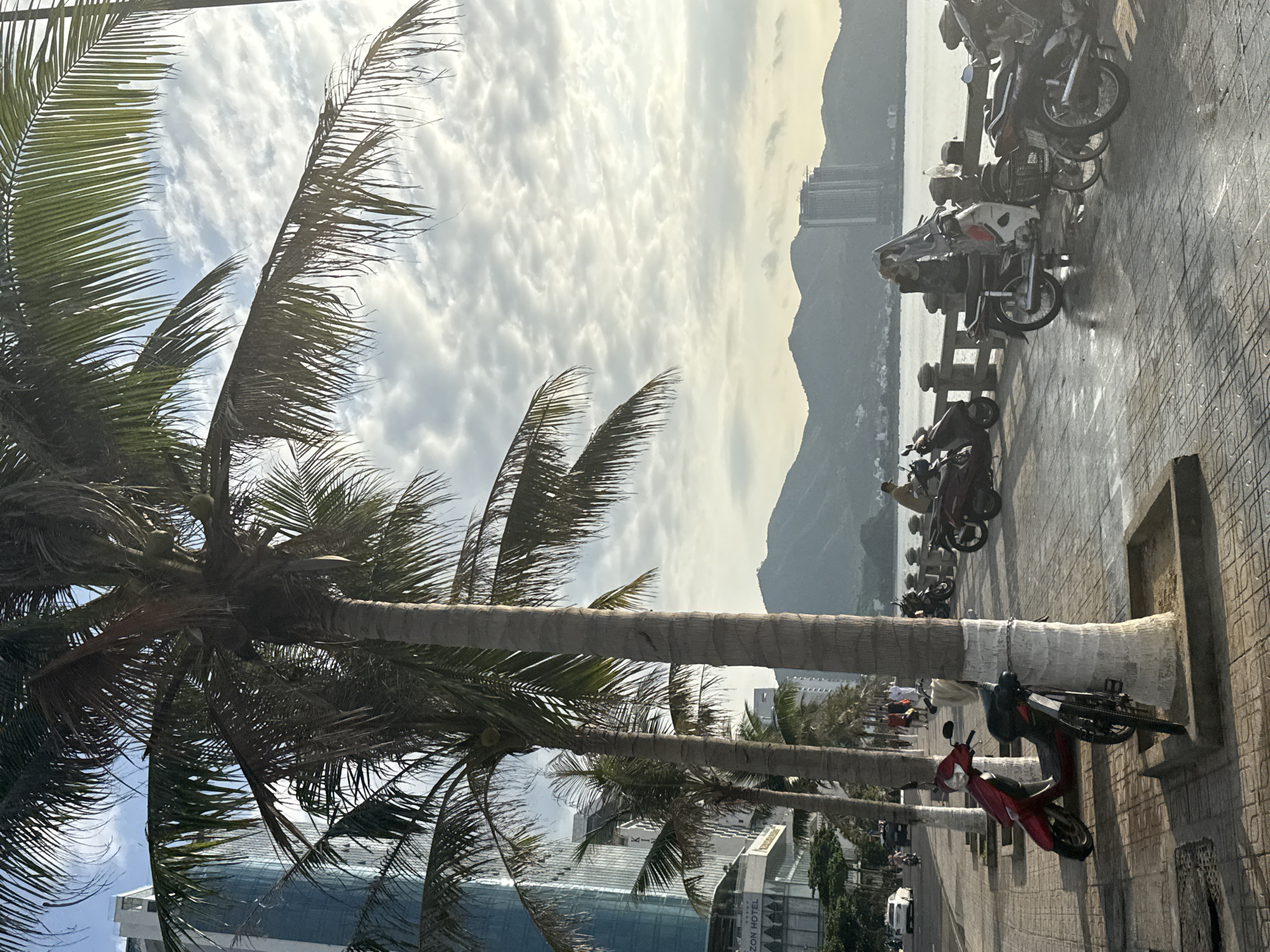 In Bac Nha Trang Ward, Vietnam, a waterfront promenade is lined with palm trees. To the left, a modern, tall blue glass building stands, with "HINZON HOTEL" visible on a lower structure. The promenade is paved with patterned tiles, and several motorcycles are parked along it, some chained to palm tree trunks. A red scooter and a bicycle are prominently placed in the foreground. In the background, a body of water stretches to a distant mountain range, with buildings visible on the far shore. The sky is filled with fluffy white clouds, and the sun casts shadows across the scene, indicating it is likely late afternoon. A few people are walking along the promenade and riding motorcycles. The atmosphere suggests a pleasant, tropical urban environment.