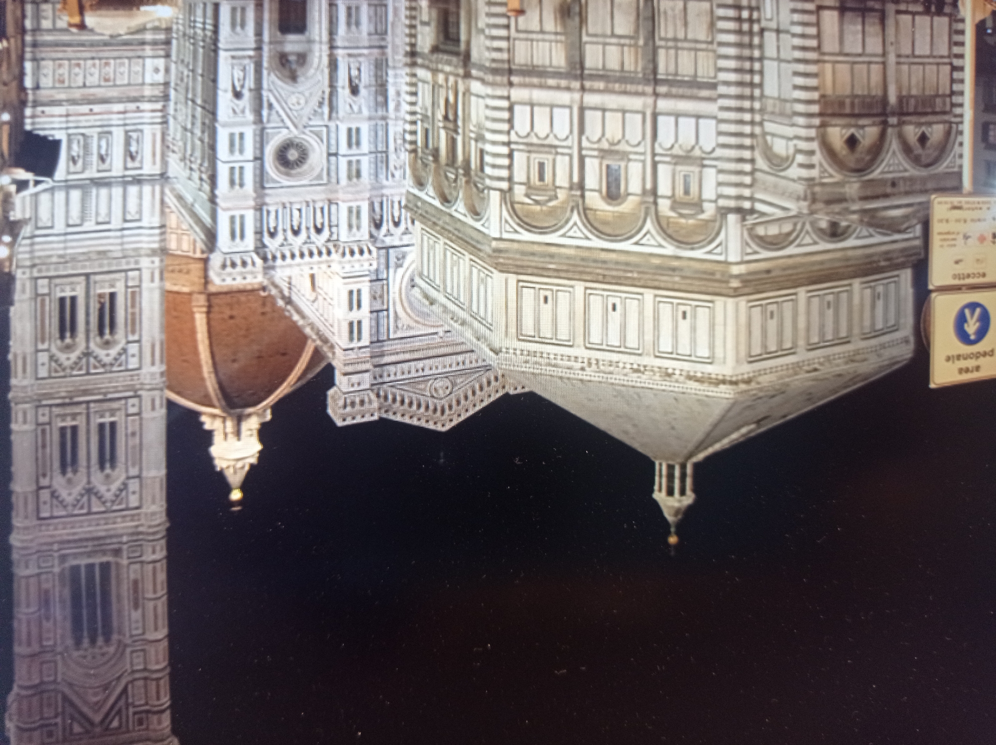The image shows a night view of the Florence Cathedral complex in Florence, Italy. The complex includes the Florence Cathedral with Brunelleschi's Dome, Giotto's Campanile, and the Florence Baptistery. The buildings are illuminated and appear to be made of white marble with decorative details. The sky is dark and filled with tiny white specks, possibly stars or dust. There are also signs visible in the foreground, one of which appears to be a pedestrian zone sign with a figure walking. The overall scene is architectural and historical. The location of Mukachevo, Ukraine is not depicted in this image.