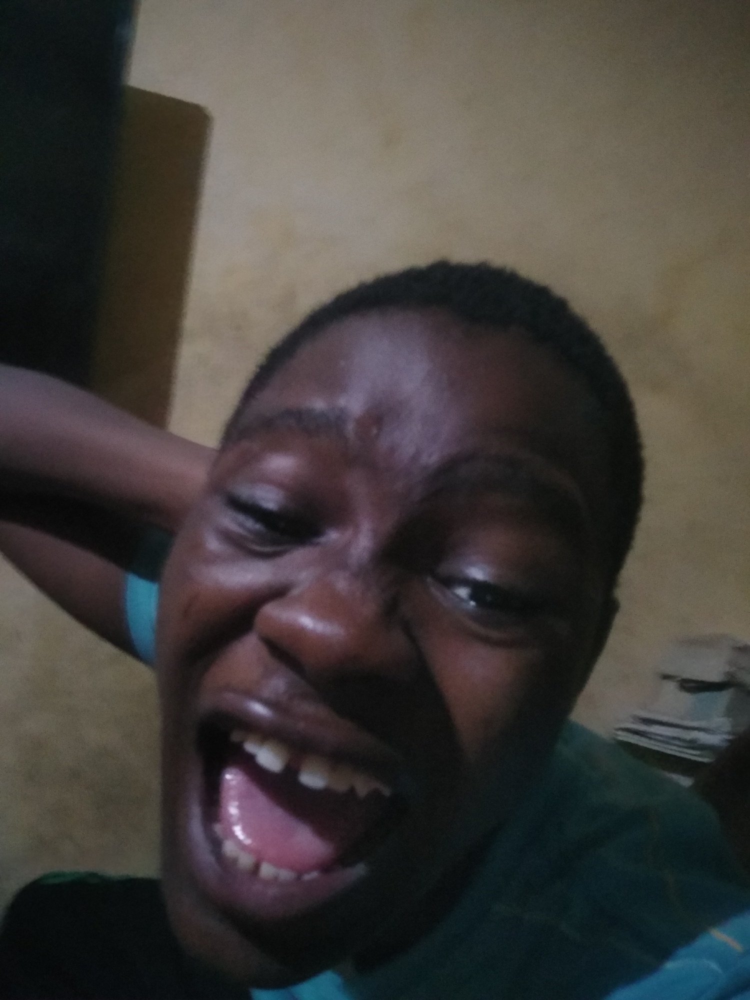 A close-up, selfie-style photograph depicts a person with dark skin, short hair, and visible teeth, mouth open wide. The person's face is the primary subject, showing expressions around the eyes and mouth. Part of an arm, possibly the person's own, is visible near the top left. In the background, a neutral-colored wall is discernible. There are some indistinct objects or papers stacked in the background, to the right of the person's head. Location information is not discernible in the image.