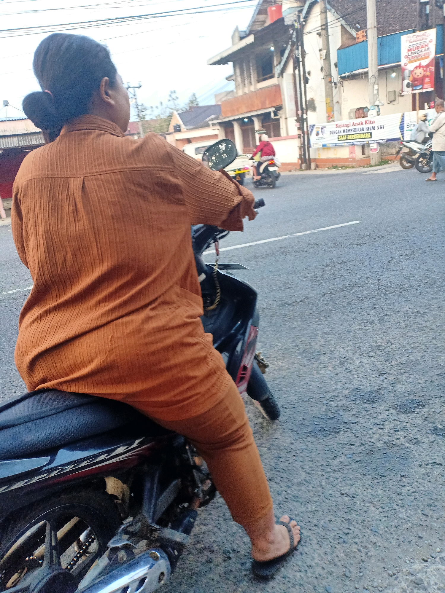 A woman, viewed from the back, is seated on a dark-colored motorcycle on a paved road. She is wearing a rust-colored, ribbed shirt and matching rust-colored pants. Her dark hair is pulled back into a bun with a small ponytail accent. The motorcycle's exhaust pipe is visible. In the background, there are buildings, a utility pole, and a street with other motorcycles and people. A banner with Indonesian text is visible on a building, and another banner with text and images is also present. The road appears to be a public thoroughfare in a city, likely Mojokerto, Indonesia, given the context.