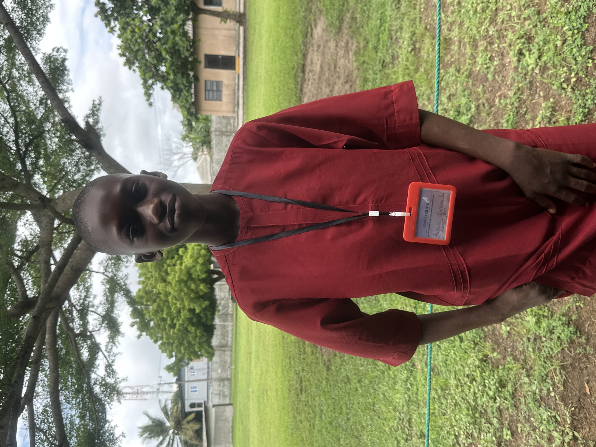 A young boy, appearing to be of Nigerian ethnicity and possibly under 18 years of age, stands outdoors in Itoga, Nigeria. He is wearing a traditional red garment, possibly an agbada, with a black lanyard around his neck. Attached to the lanyard is a bright orange identification badge holder containing a white card with handwritten text in black ink, some of which is legible. The boy's hands are tucked into the front pockets of his garment, and he is looking directly at the camera with a neutral expression. He has short hair and dark skin.
The background consists of a grassy field, a mature tree with an expansive canopy, and in the distance, buildings and a cell tower. The sky is overcast. The scene suggests a casual outdoor setting, potentially within a school or community compound, given the presence of the ID badge.