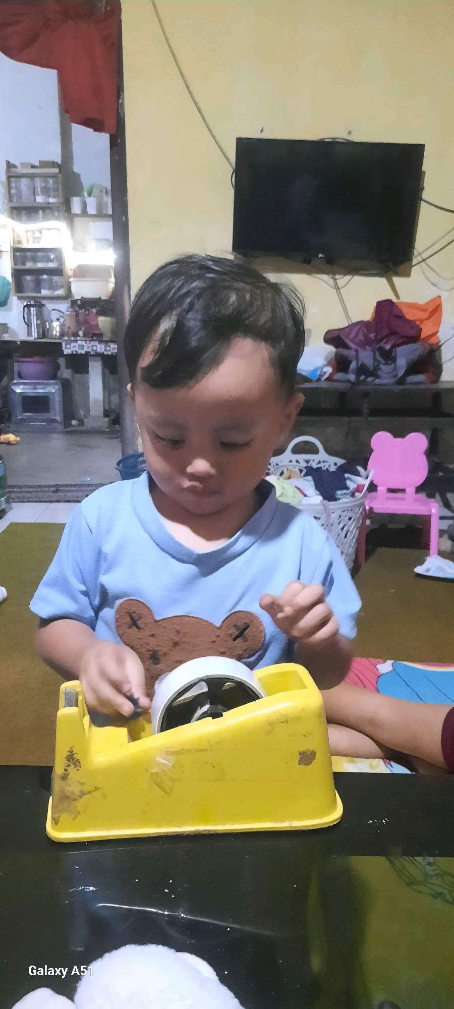 A young child, likely male, wearing a light blue t-shirt with a brown bear graphic is seated at a black table in a room. The child is interacting with a yellow toy tape dispenser. The background of the room shows a yellow wall, a dark television mounted on the wall, shelves with various items, and a pink child-sized chair. The environment appears to be a domestic setting. The image metadata indicates the location is Jatitengah, Indonesia.