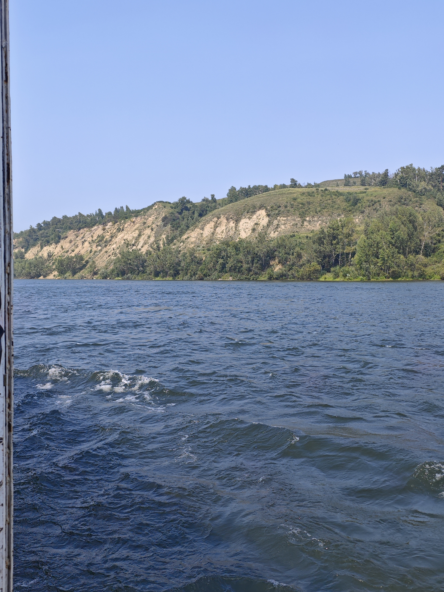 The image depicts a wide, calm river with gentle waves rippling across its surface. The water is a deep blue color. On the far bank of the river, a steep, eroded hillside rises, displaying layers of light brown soil and rock. This hillside is partially covered with green vegetation and scattered trees. The sky above is a clear, pale blue. A portion of a weathered, metallic structure, possibly a railing or part of a boat, is visible on the left edge of the frame, adding a sense of being on the water. The scene appears to be set in a natural, undeveloped environment. While the prompt mentions the city of Khudonogovo, Russia, there are no specific landmarks or indicators in the image to confirm this exact location.