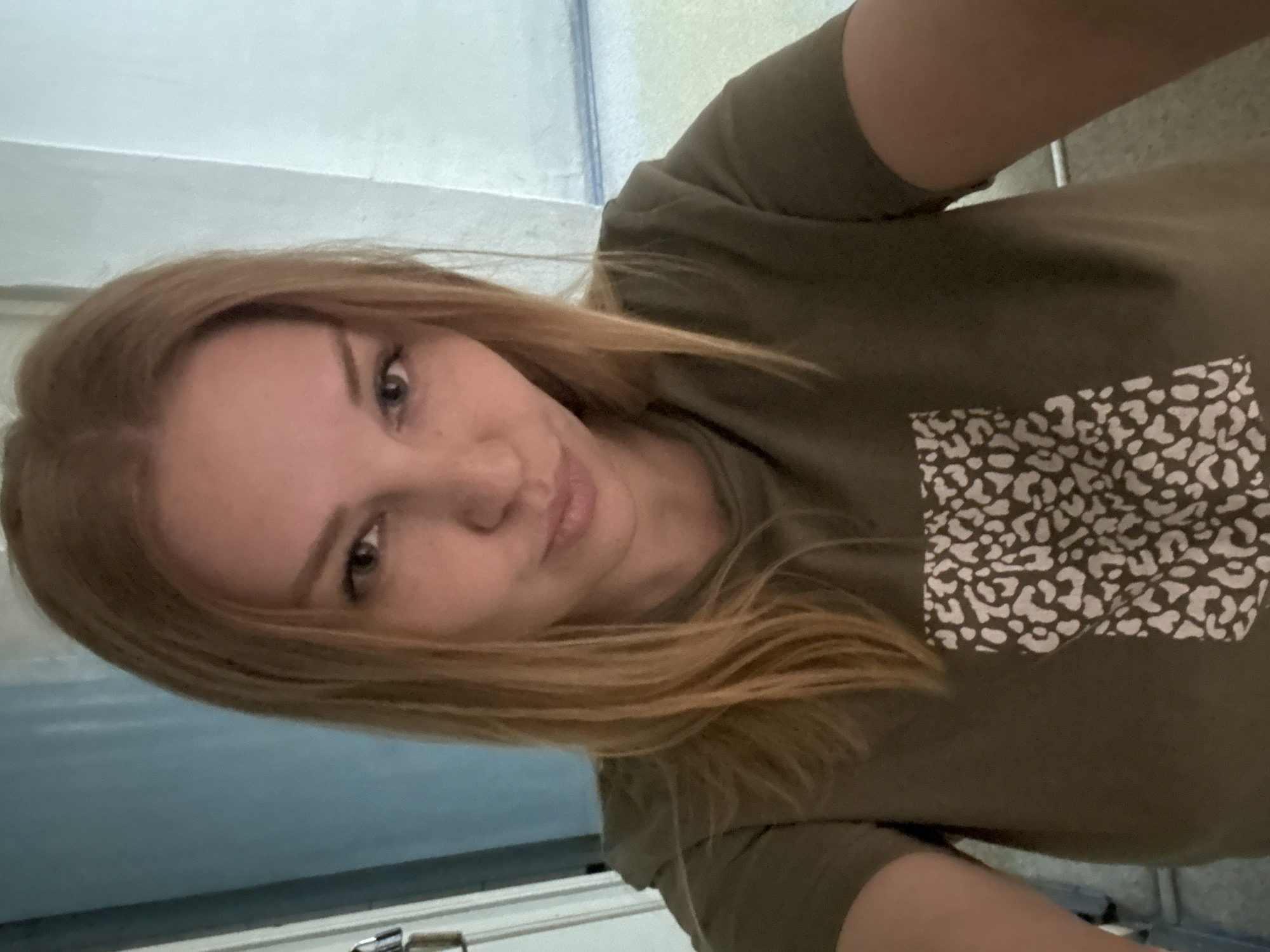 The media file is a self-portrait of a young woman with blonde hair, taken indoors. She is wearing a dark green t-shirt with a white and dark patterned pocket. The background appears to be a domestic setting, with what looks like a light-colored wall and possibly a doorway or cabinet. The lighting suggests it is daytime, and the environment seems calm and personal. The woman's expression is neutral, and the image is a close-up of her face and upper body, with the camera angled slightly from above. There is no visible text in the image. The provided location context of Заволжье, Russia, suggests the setting is within that region.