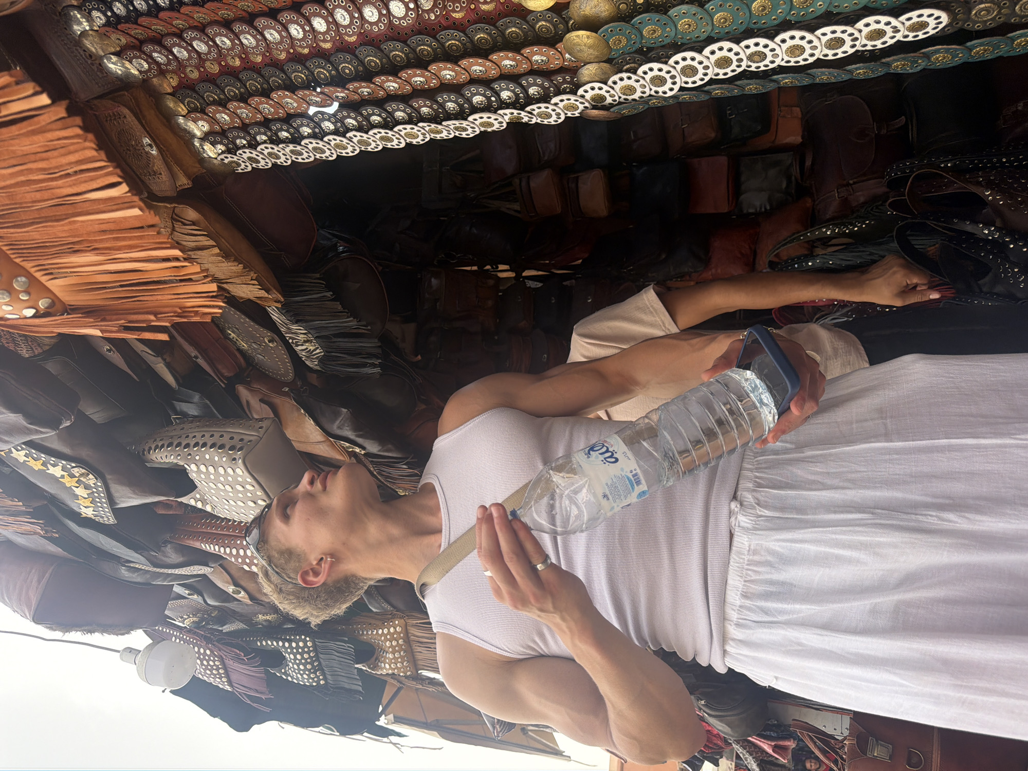 The media file shows a person holding a water bottle in a marketplace setting. The person is wearing a white tank top and white pants. They have short blonde hair and are looking to their left. The marketplace appears to be a stall selling leather goods, with many bags and accessories on display. The items are adorned with decorative metal studs and fringe. The stall is illuminated by a light bulb hanging from the ceiling. The overall atmosphere is that of a busy market. The image is shot outdoors, and the lighting suggests it is daytime. There is text on the water bottle in Arabic script. The location context is Marrakesh, Morocco, which is consistent with the style of goods and the likely setting of a souk.