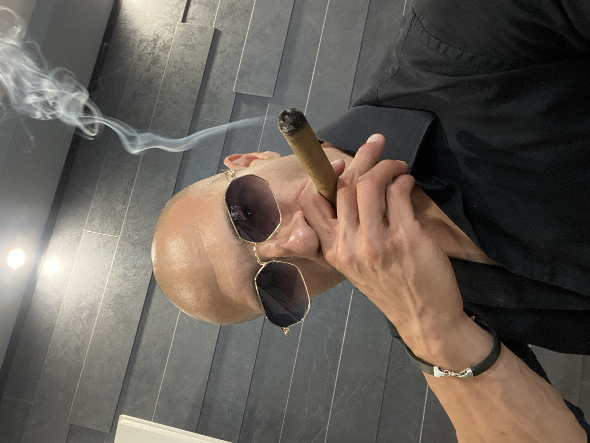 A close-up, medium shot shows a bald man wearing sunglasses and a black shirt, holding a lit cigar to his mouth. Smoke billows from the cigar, rising towards the top of the frame. The man's left arm, adorned with a black leather bracelet with silver accents, is visible in the lower left corner. The background consists of a textured, grey tiled wall. The scene is likely indoors, possibly in a residential or private setting, given the intimate framing and the man's casual attire. No specific location within Pleven, Bulgaria can be determined from the visual information provided.