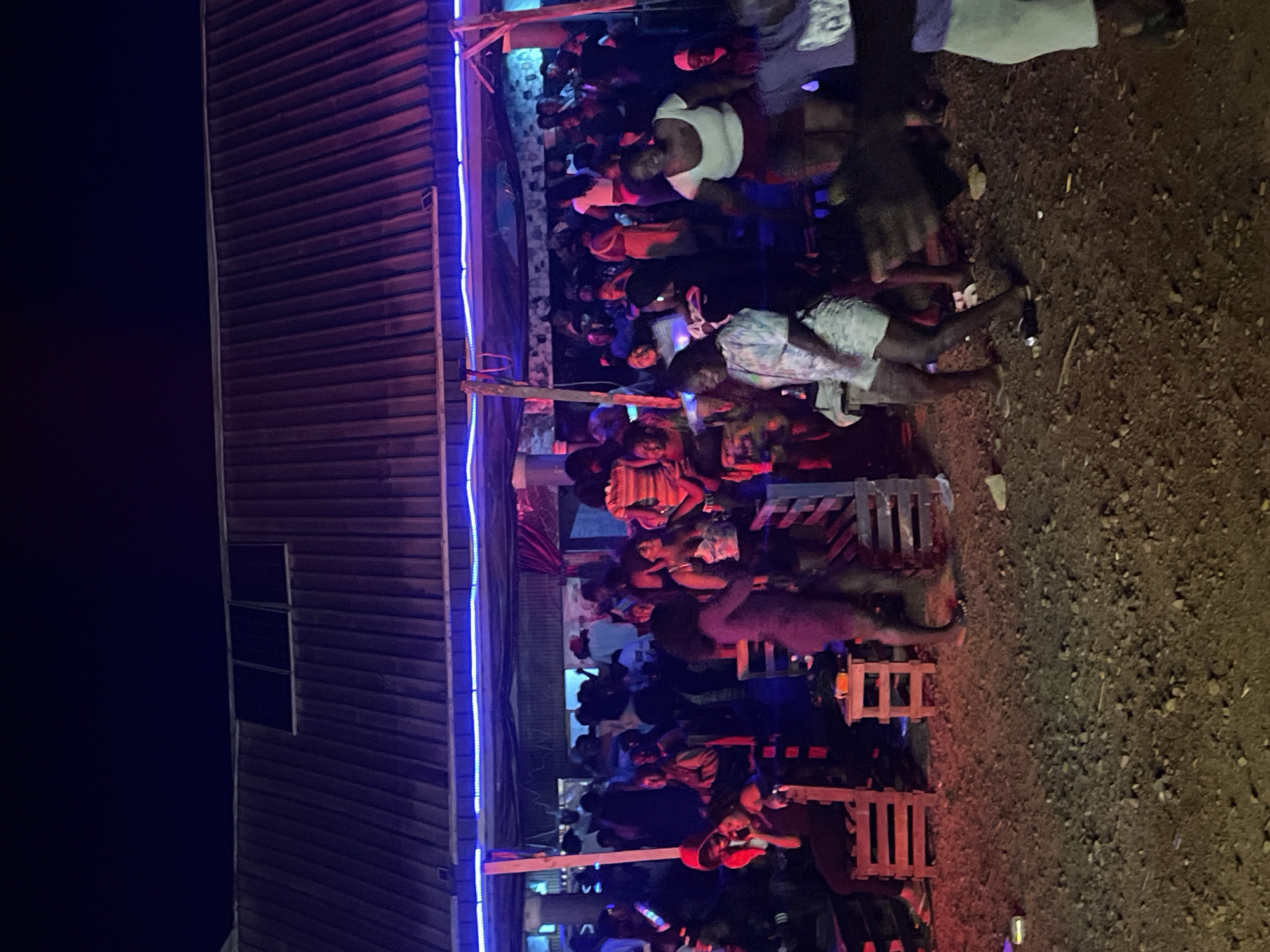 The image depicts an outdoor gathering at night, possibly a party or event, under a corrugated metal roof structure. Blue and pink lighting illuminates the scene, casting colorful reflections on the ground. A crowd of people, predominantly young adults, are seen standing and dancing. Some individuals are seated on wooden chairs and crates. The ground is covered with dirt and small stones. In the background, the structure appears to be a makeshift or open-air venue. The location is identified as Durumi, Nigeria.
