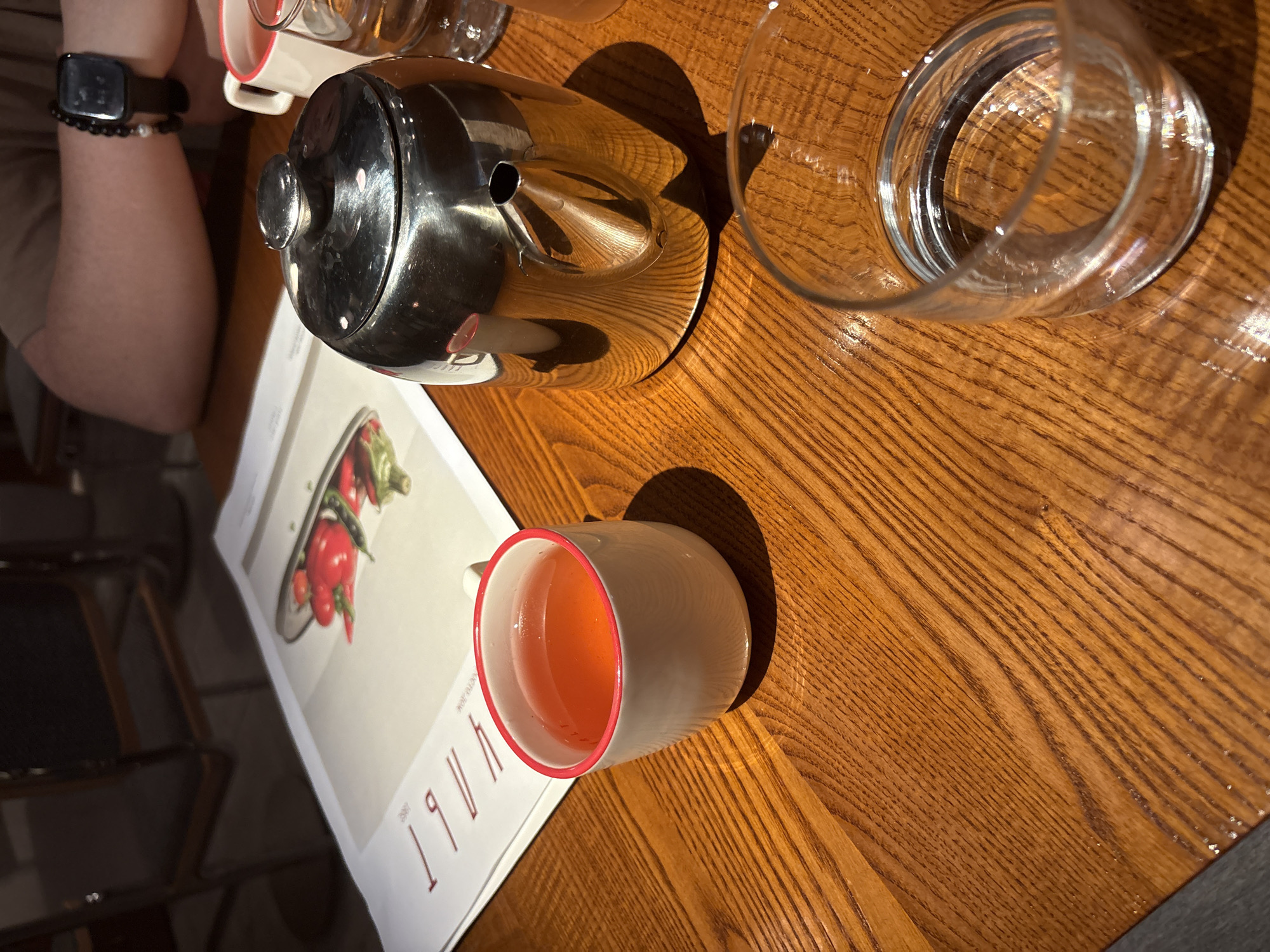 A close-up shot of a wooden table shows a tea set and a menu. On the left, a white teacup with a red rim contains a red beverage. Next to it, a shiny metal teapot with a silver lid and handle is positioned on the table. To the right of the teapot, a clear glass of water is visible. In the background, a person's arm with a black beaded bracelet and a smartwatch is resting on the table. A menu with an image of red vegetables is also placed on the table. The scene appears to be set in a restaurant or cafe in Rostov-on-Don, Russia, based on the provided context.