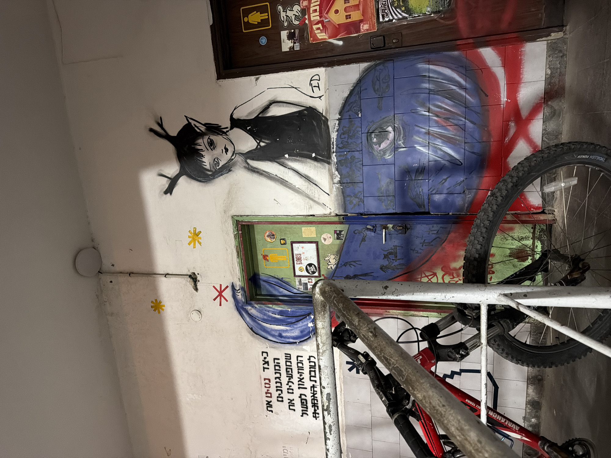 An indoor hallway or stairwell in Tel-Aviv, Israel, features a vibrant display of street art and everyday objects. The dominant subject is a large, black and white graffiti drawing of a stylized female character with large eyes and spiky hair, wearing a dark top, painted on a light-colored wall. The letters "ID" are visible next to her.
Below the character, a portion of a blue mural with abstract designs, possibly depicting a face, extends across the wall and partially onto a lower door. To the right of this, red and white stripes form a pattern, including an 'X' shape. An upper, dark brown door is adorned with various stickers and posters, including a yellow unisex restroom symbol and a Hebrew poster reading "נוכת בית" (House Warming/Homecoming). A second, green door is visible below the murals, also covered in stickers.
To the left of the lower door, blue, fabric-like shapes are painted on the wall. Below these, a block of Hebrew text reads, "כיצד נטיט את המנגנונינו שמפעילינז את המוזיאון לפעול לטובת האמניו*" (How to steer our mechanisms that activate the museum to act for the benefit of the artists*). Three star-shaped decals or paintings—two yellow and one red—are placed further left on the wall.
Two bicycles are parked along a metal railing in the foreground, partially obscuring the right side of the wall art. One red bicycle has "AVALANCHE ALL TERRA" visible on its frame, and a tire with "KENDA NEVEGAL" tread is prominent. The setting is brightly lit, likely by artificial light, given the indoor context. No specific activity is taking place beyond the static arrangement of art and parked bikes.