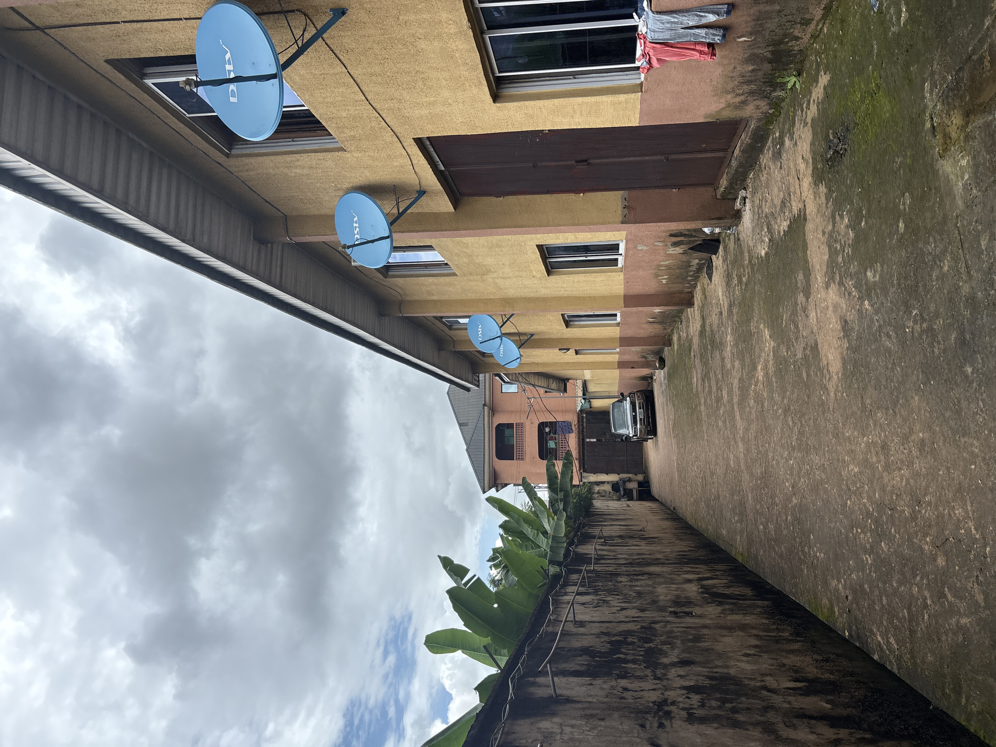 This is an outdoor, eye-level, medium shot of an alleyway in Onitsha, Nigeria, with buildings on the right and a concrete wall with large banana leaves on the left. The sky is overcast with grey clouds. The building on the right is a two-story structure painted yellow and rust brown, with several windows and satellite dishes mounted on the exterior. Clothes are hanging on a clothesline outside one of the windows. The alleyway is paved with concrete and has some moss and puddles on the ground. A dark-colored car is parked in the distance. The wall on the left is weathered and covered in dark stains, with metal rods protruding from the top. In the background, another building with balconies is visible. There are no people visible in the image.